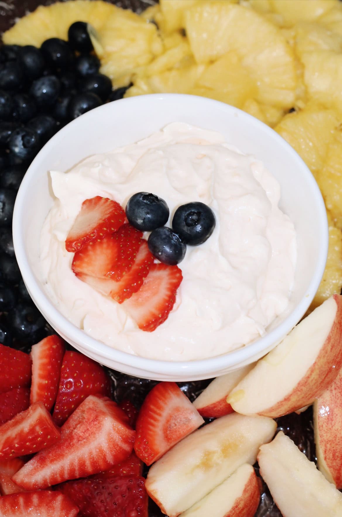 White bowl of fruit dip with strawberries and blueberries surrounded by apples, pineapple, blueberries, and strawberries