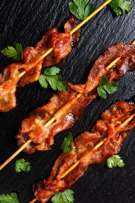 Candied bacon skewered on wooden sticks lies on a black grained surface.