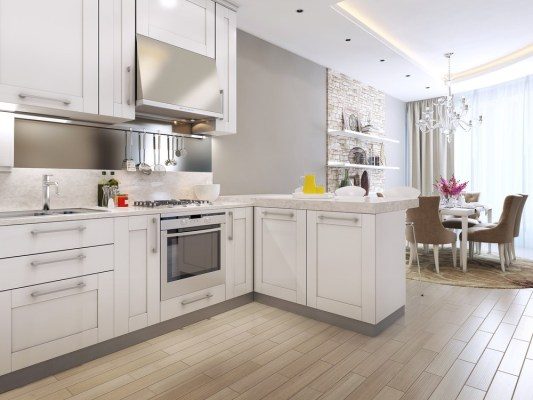Modern kitchen with white cabinets, stainless steel appliances, and light wood flooring. The open layout makes it easy for a Roomba to keep your house clean easier while you enjoy meals in the dining area with a round table, chandelier, and large window.