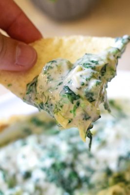 Tortilla chip dipped in 3 ingredient spinach and artichoke dip being held by a non-descript hand