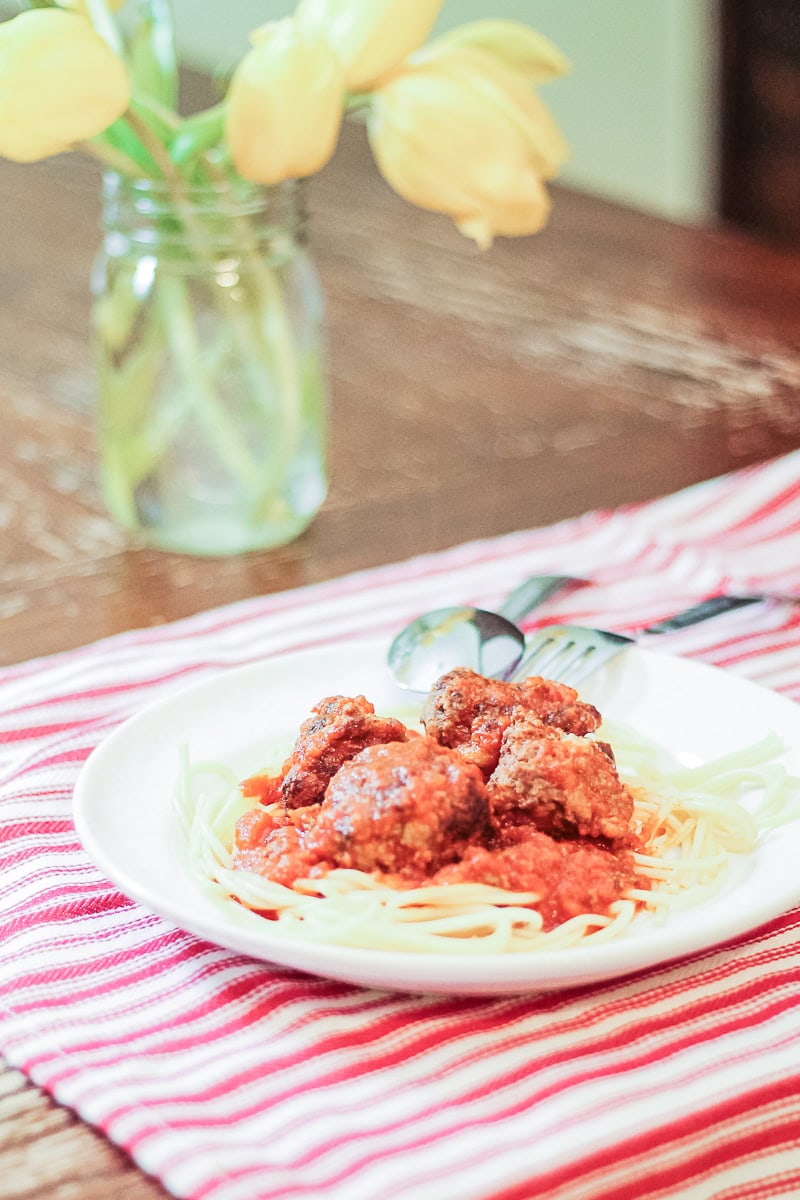 spaghetti and meatballs on a white plate with a fork and spoon and yellow tulips in the background
