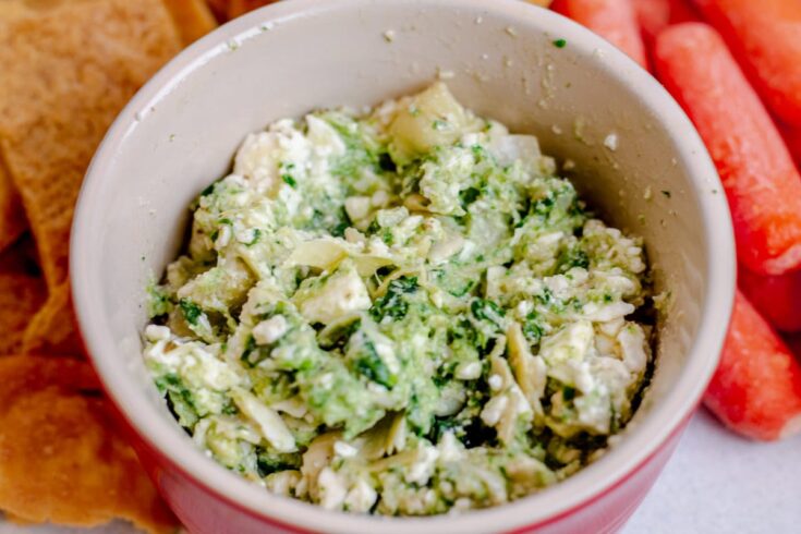 Red bowl with spinach, artichoke, and feta Mediterranean appetizer dip with pita chips and carrots