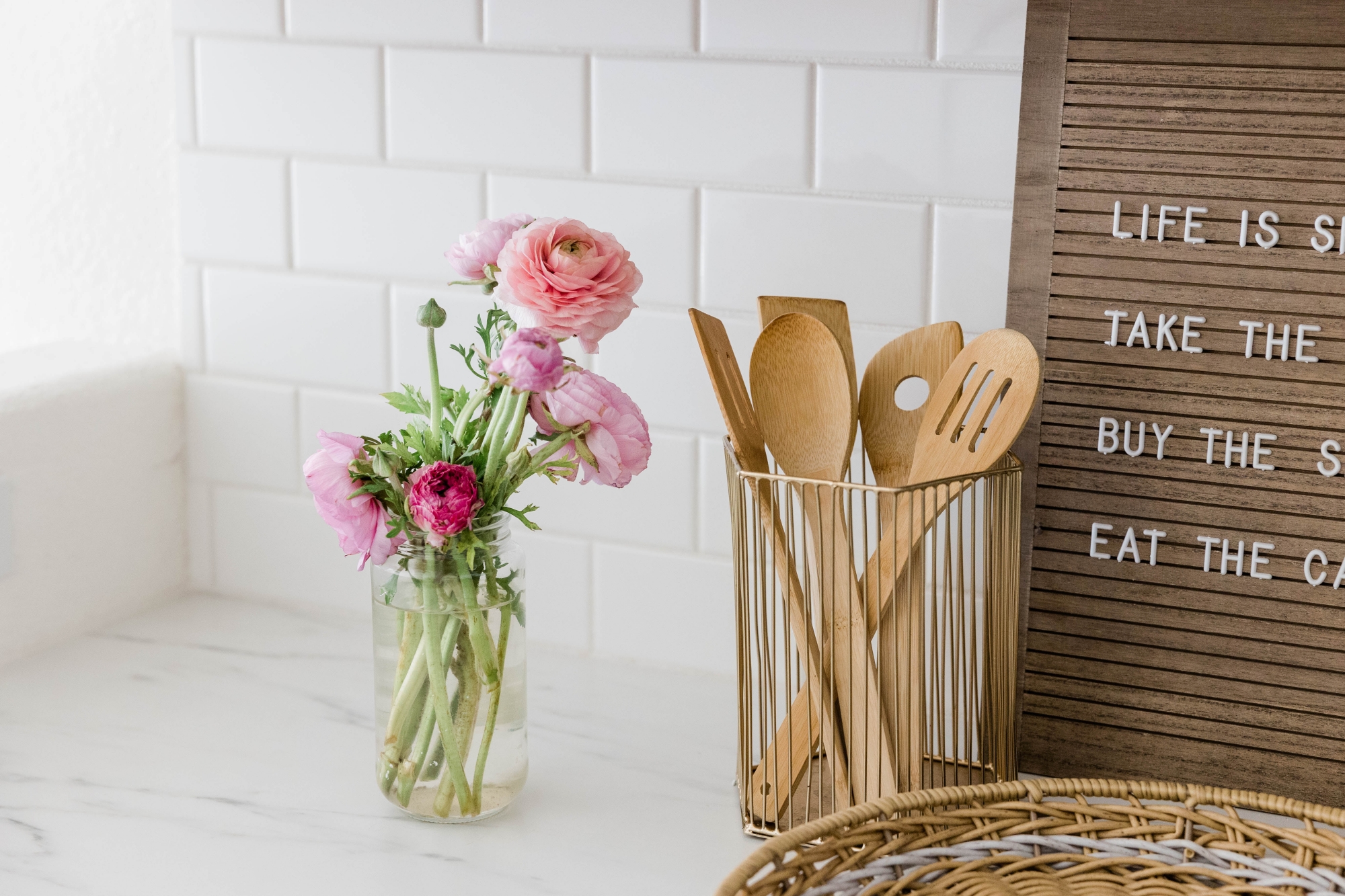 Circle cropped photo of fresh pink carnations and a wire canister of wooden kitchen utensils on a kitchen counter.