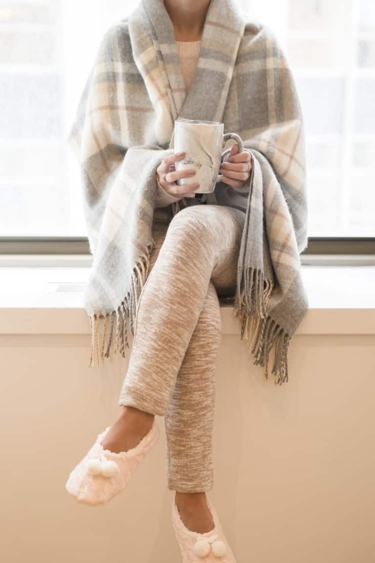 non descript woman sitting on a counter while holding a coffee mug and wearing a shawl, leggings, and slippers