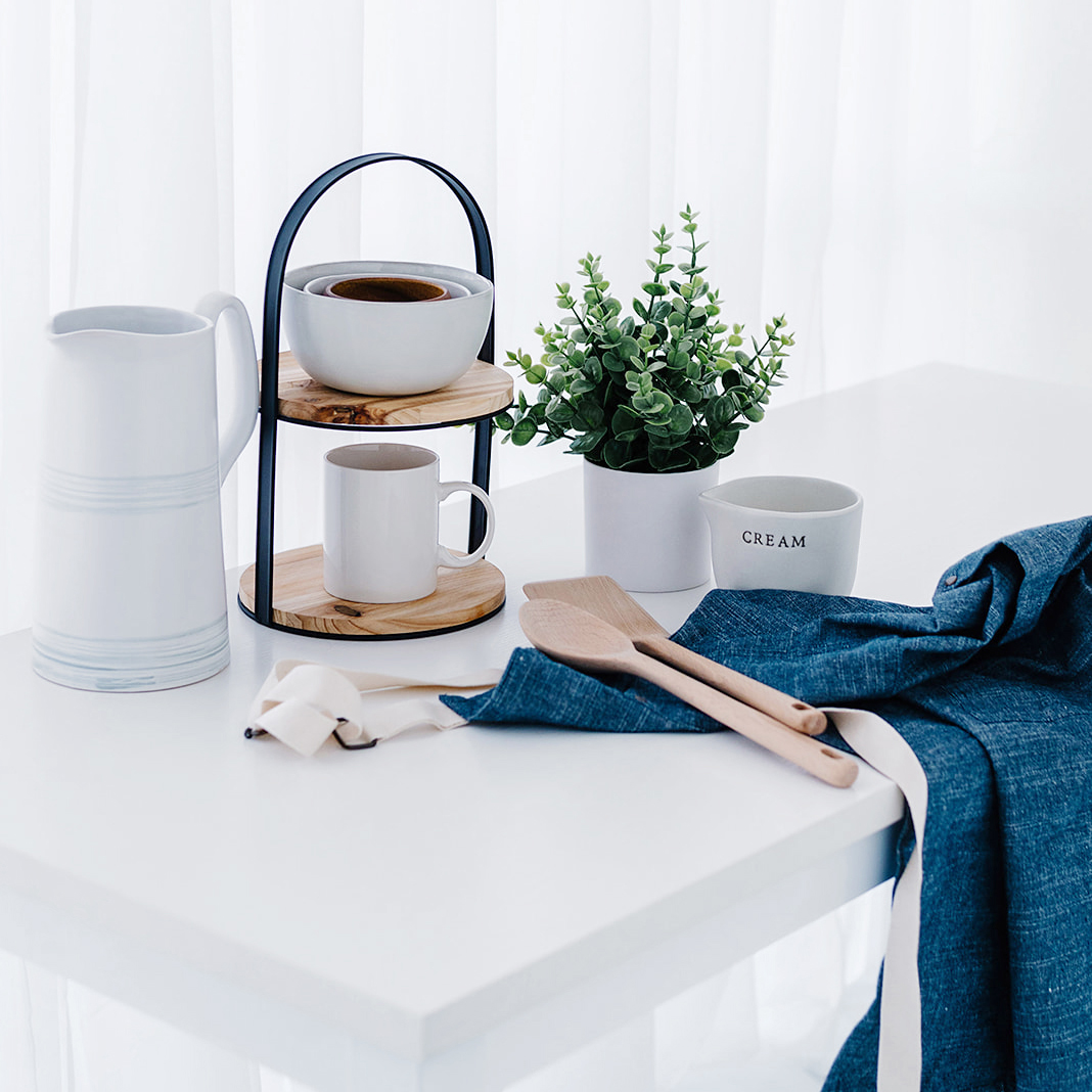 minimalist kitchen with pitcher, tiered cake stand, mixing bowls, coffee mug, wooden spoons, faux plant, and denim apron