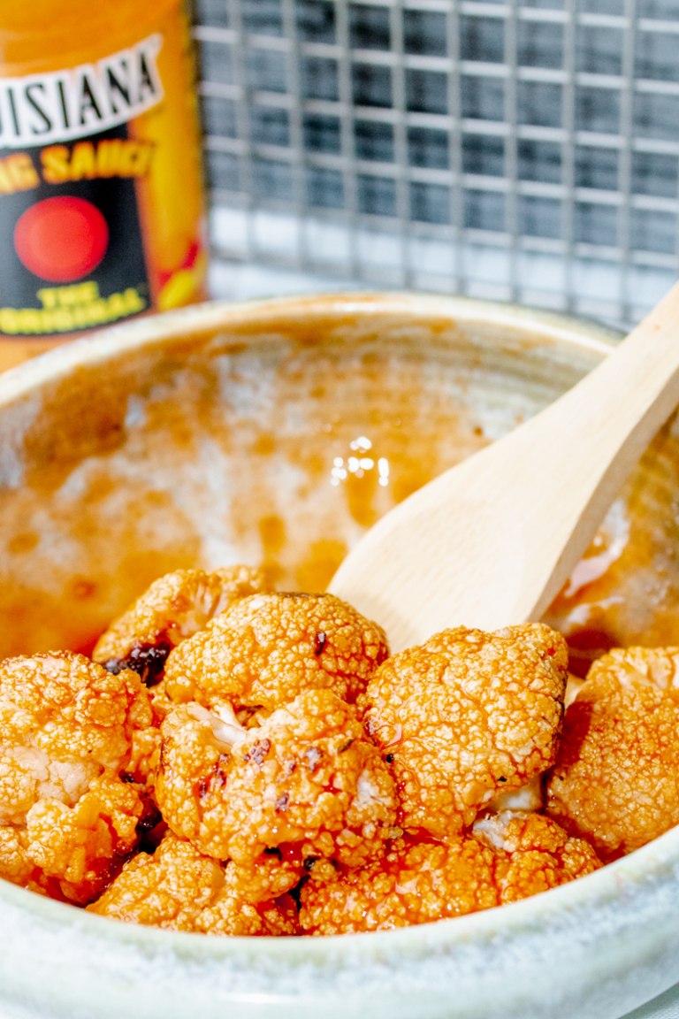 roasted buffalo cauliflower bites in a bowl with a wooden spoon and hot sauce in the background