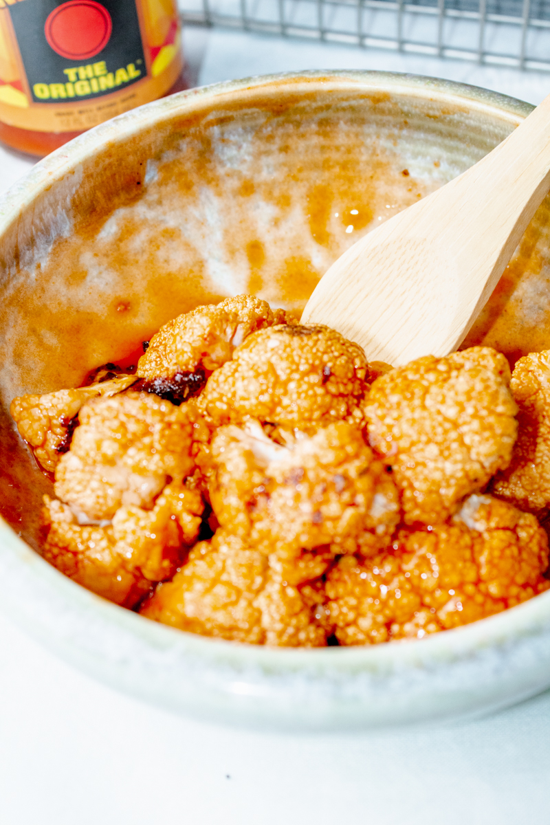 close up of roasted buffalo cauliflower bites in a bowl with a wooden spoon and hot sauce in the background