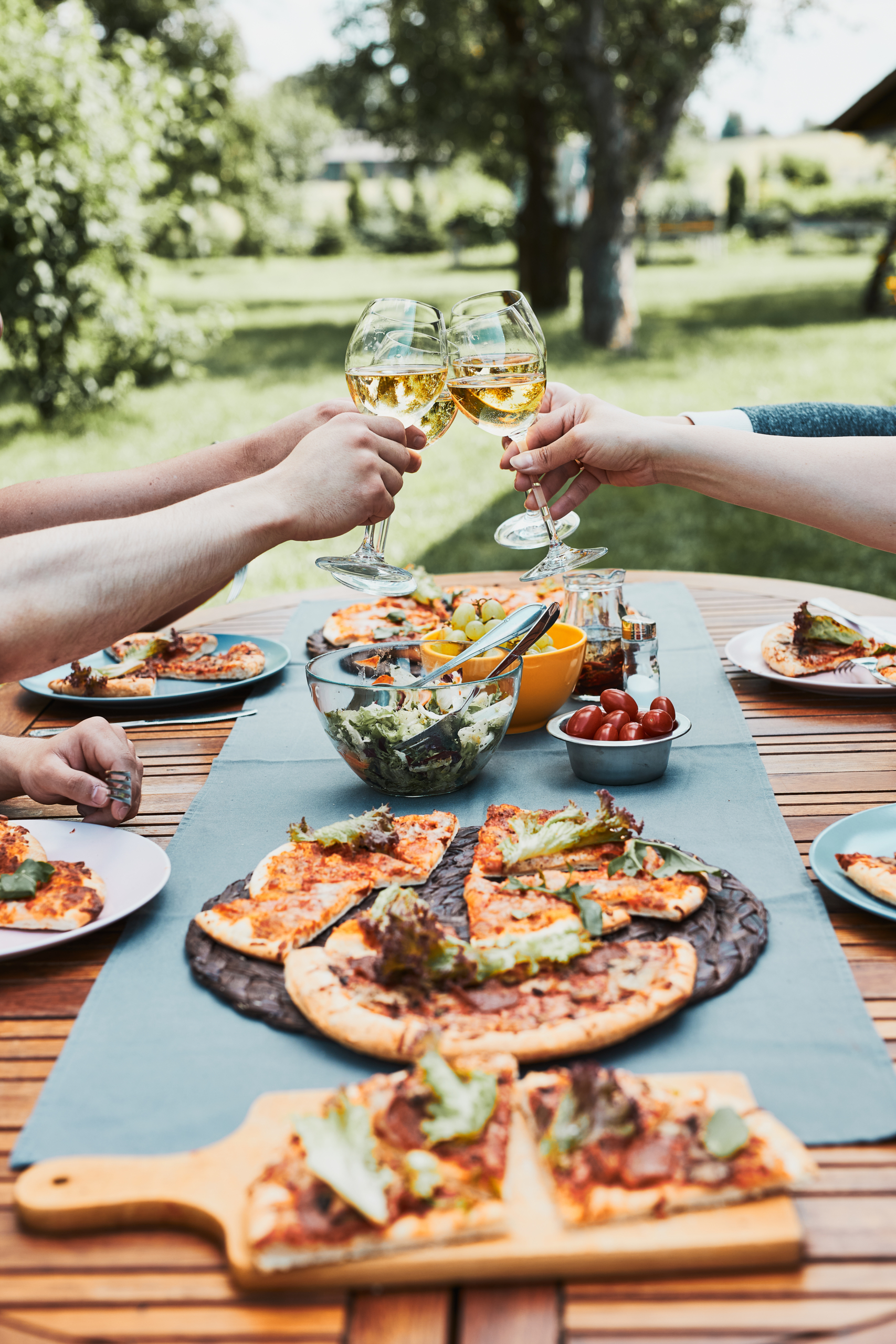 Friends making toast during summer picnic outdoor dinner in a home garden. Close up of people holding wine glasses with white wine over table with pizza, salads and fruits. Dinner in a orchard in a backyard