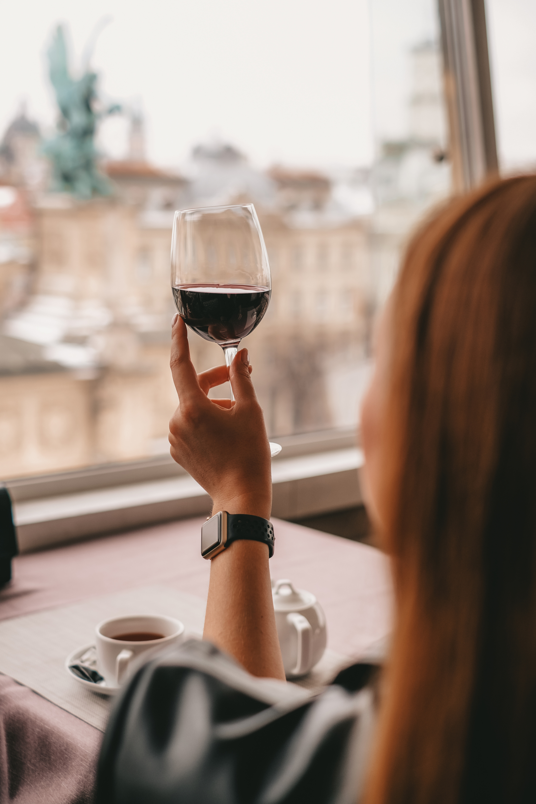 woman sitting alone at a table, looking out the window, holding up a glass of red wine