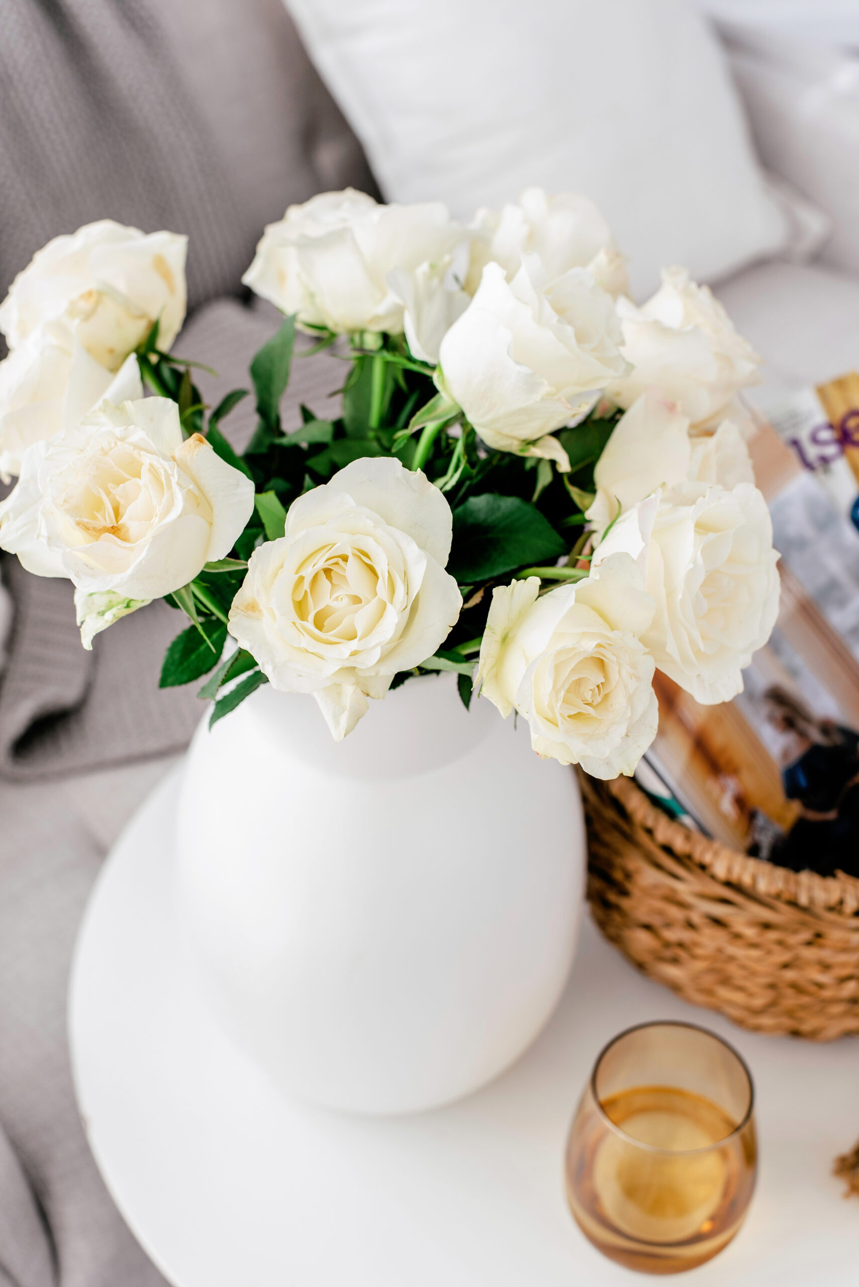 overhead view of a vase with white roses on a table with a glass of wine and a basket of magazines