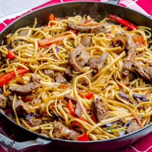 Black pan on a red checkered cloth filled with beef, noodles, peppers, and cabbage that has been stir fried