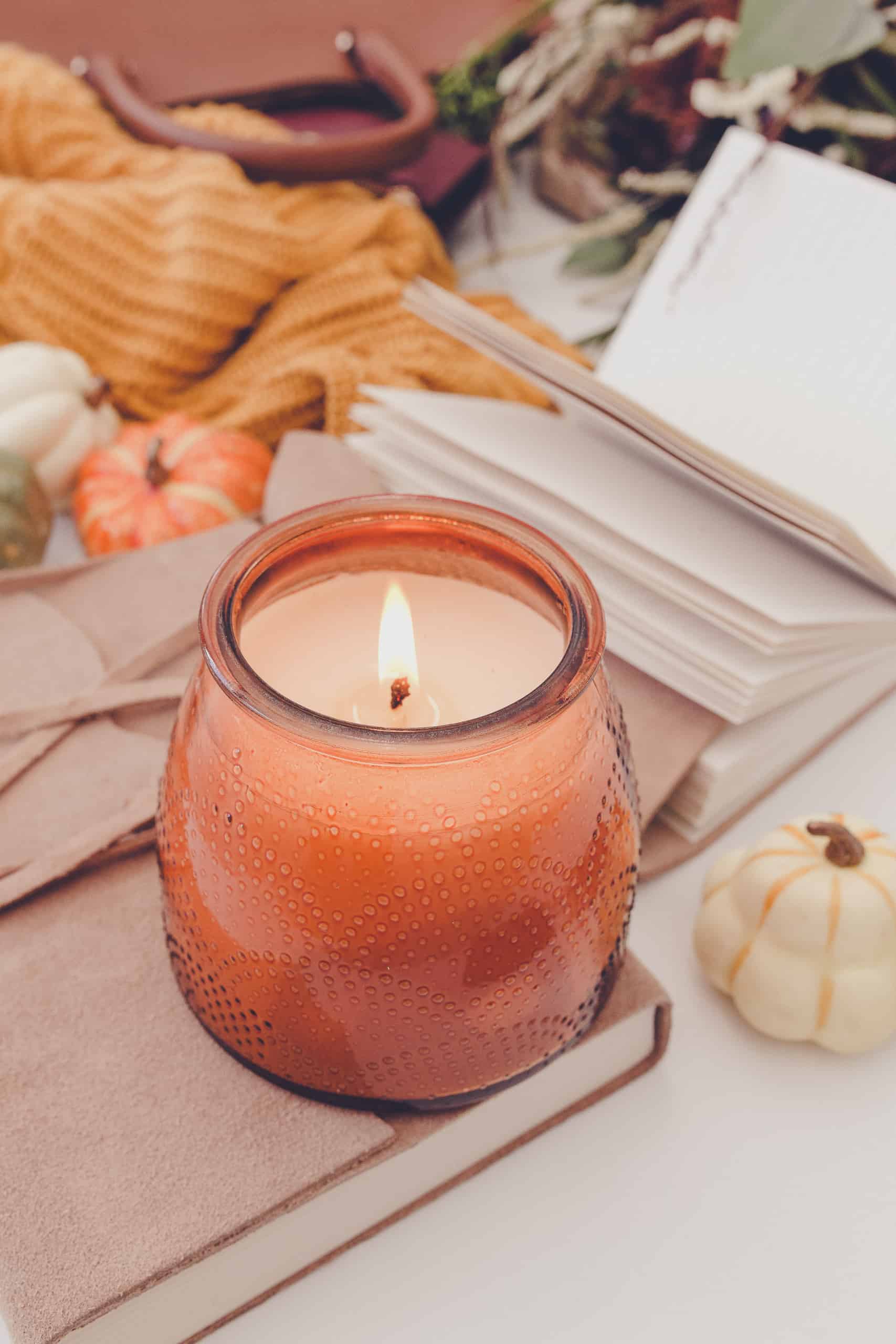 glass container with a burning candle surrounded by cozy fall pumpkins, a blanket, and a book