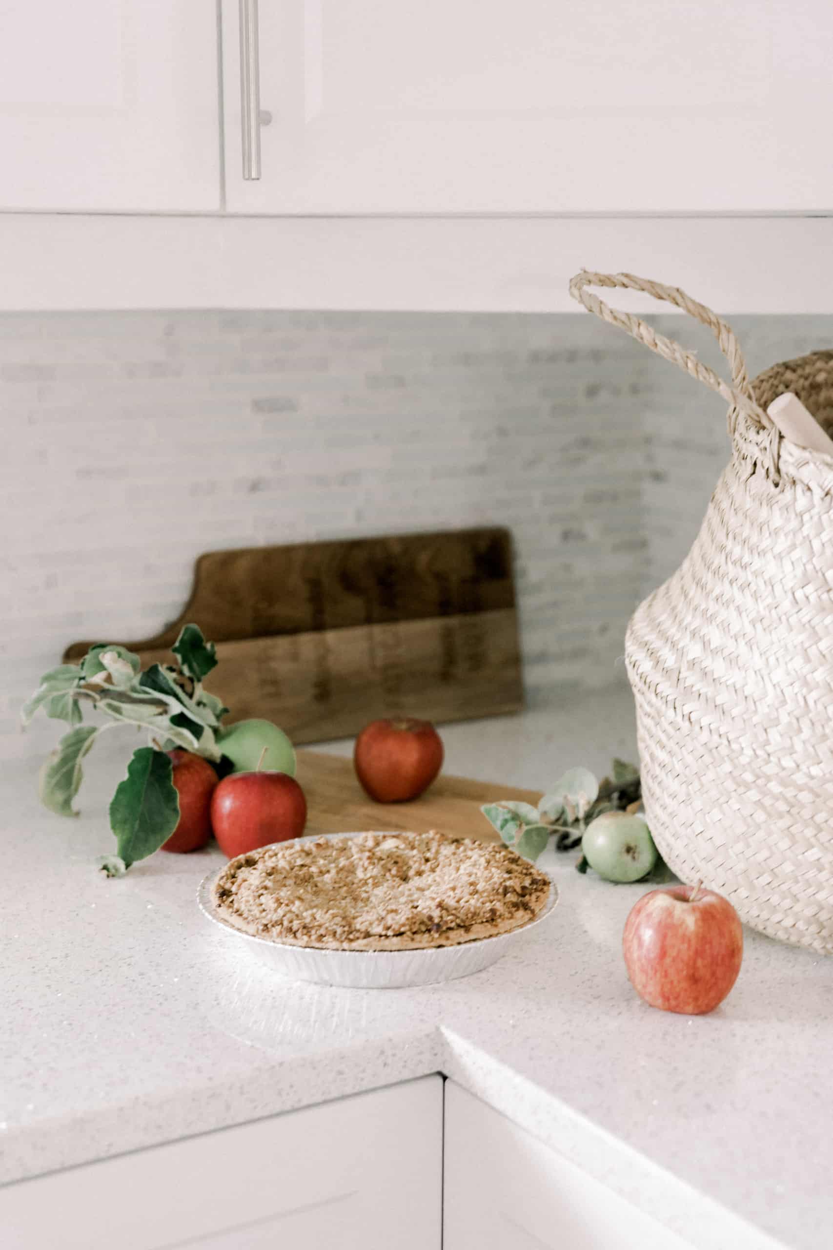 view of a kitchen counter with an apple pie, apples strewn about, a wooden cutting board in he background, and a market basket