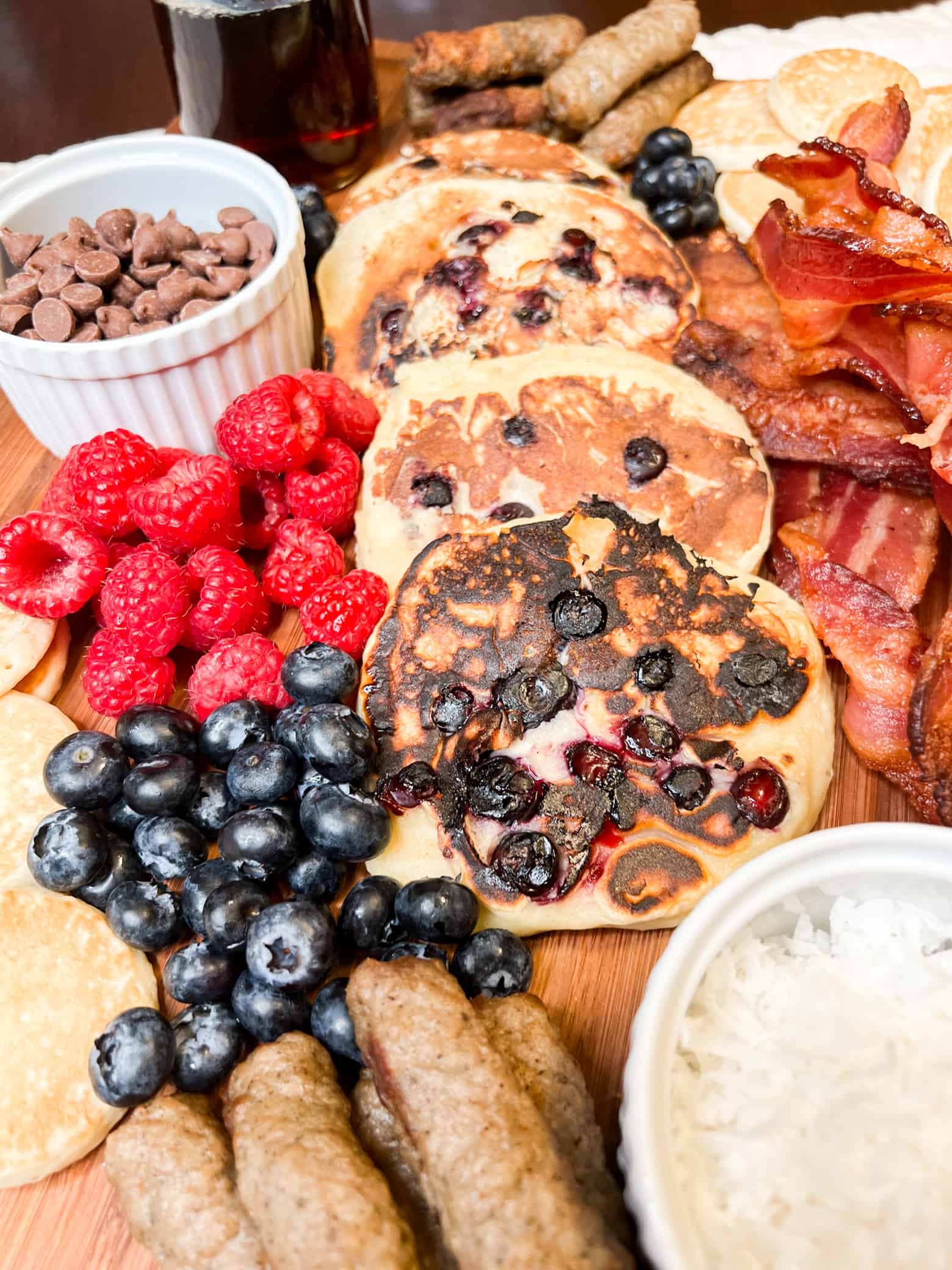 close up of a charcuterie board of breakfast items including pancakes, sausage. bacon, berries, and chocolate chips