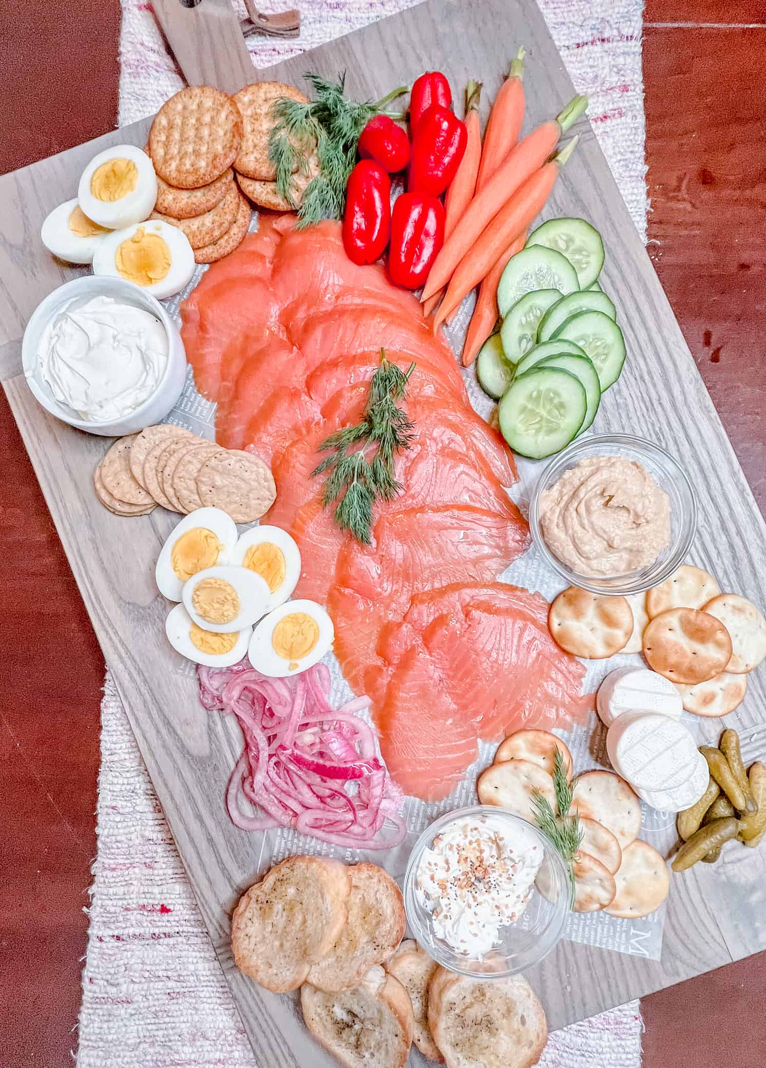 overhead view of smoked salmon charcuterie with fresh veggies, asorted crackers, hard-boiled eggs, humus, cream cheese, brie, and pickled red onions