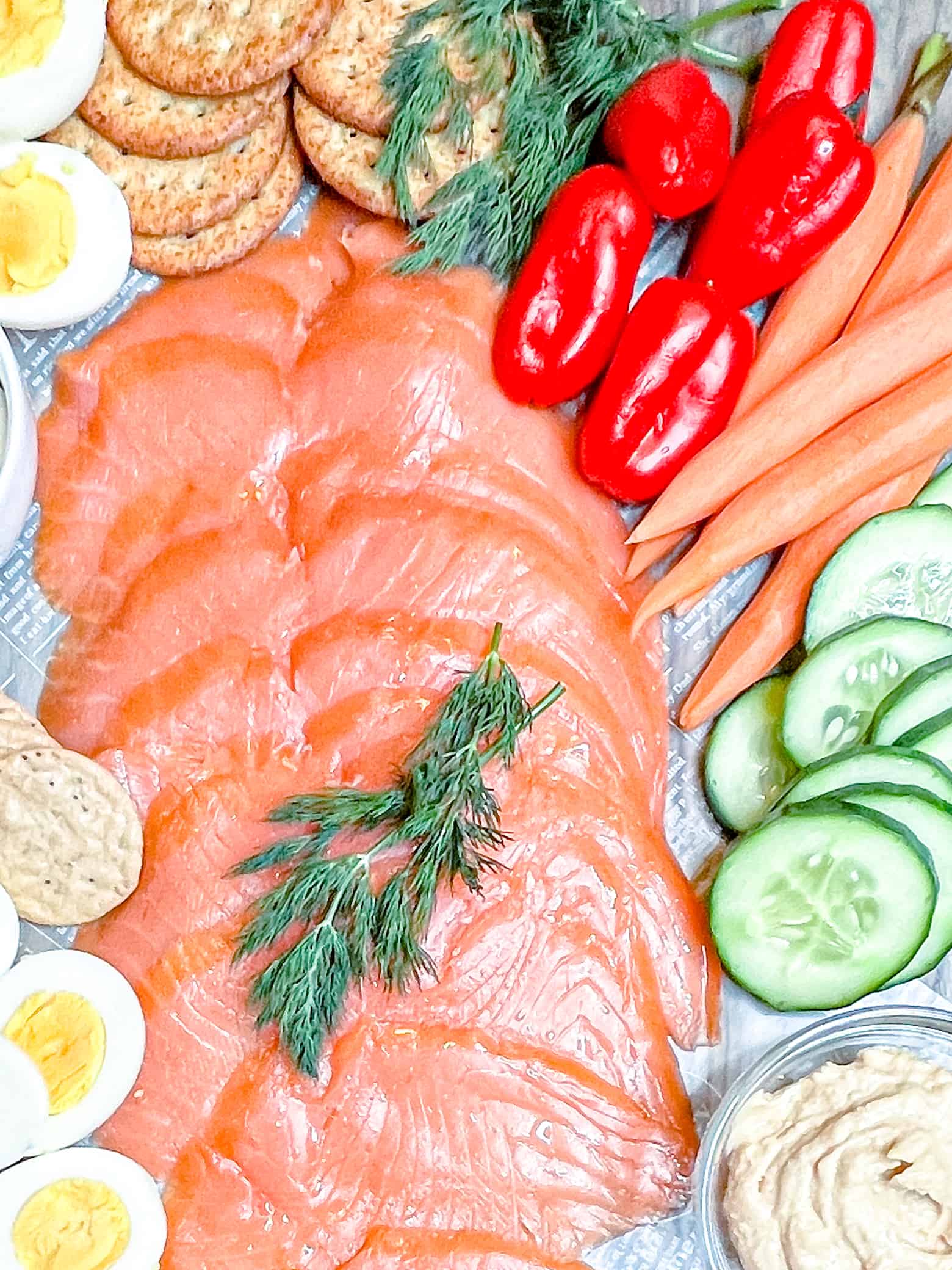 overhead view of smoked salmon charcuterie with fresh veggies, asorted crackers, hard-boiled eggs, and dill