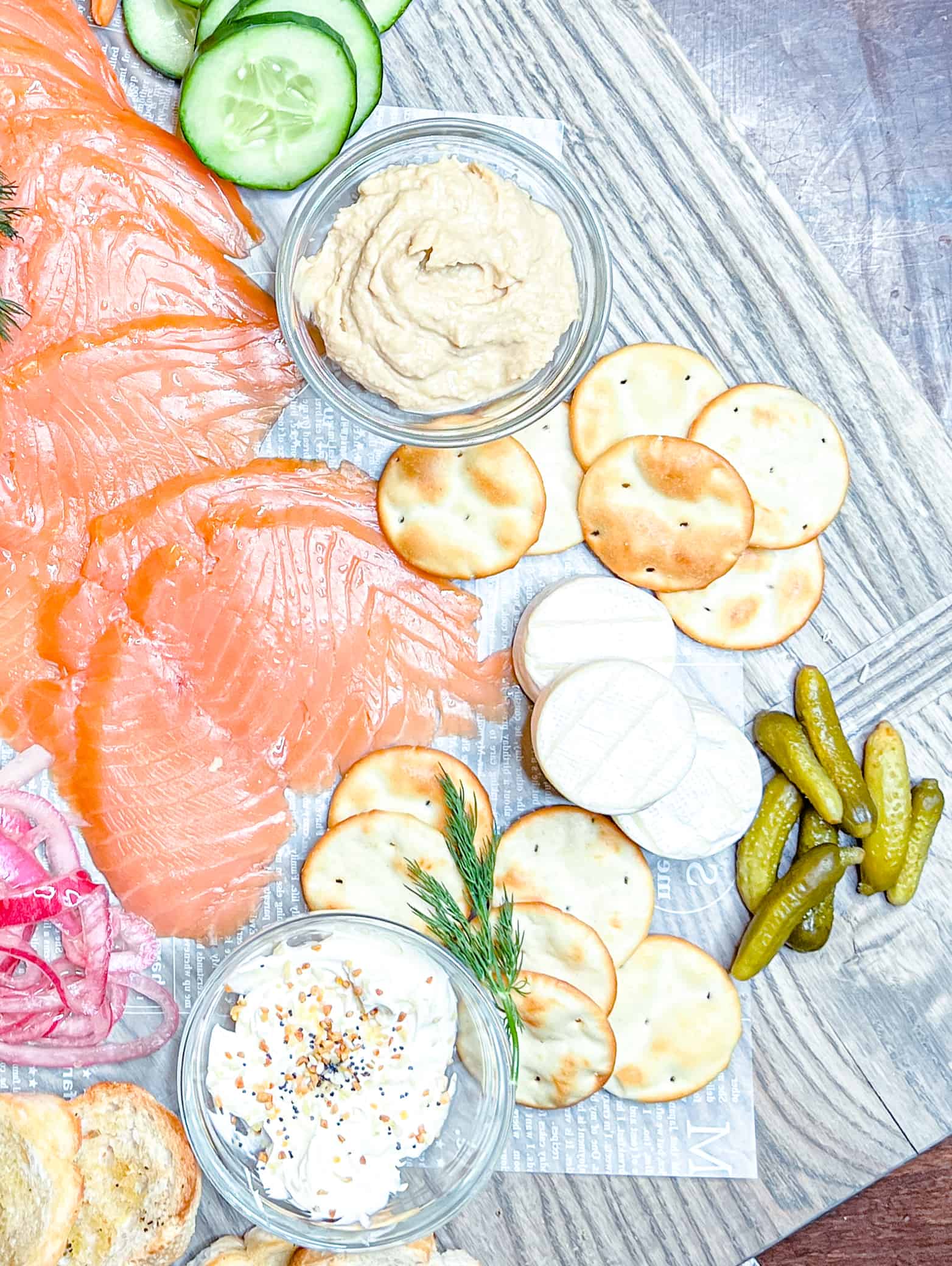 overhead view of a charculterie board with smoked salmon, humus, cream cheese, dill, pickles, and crackers