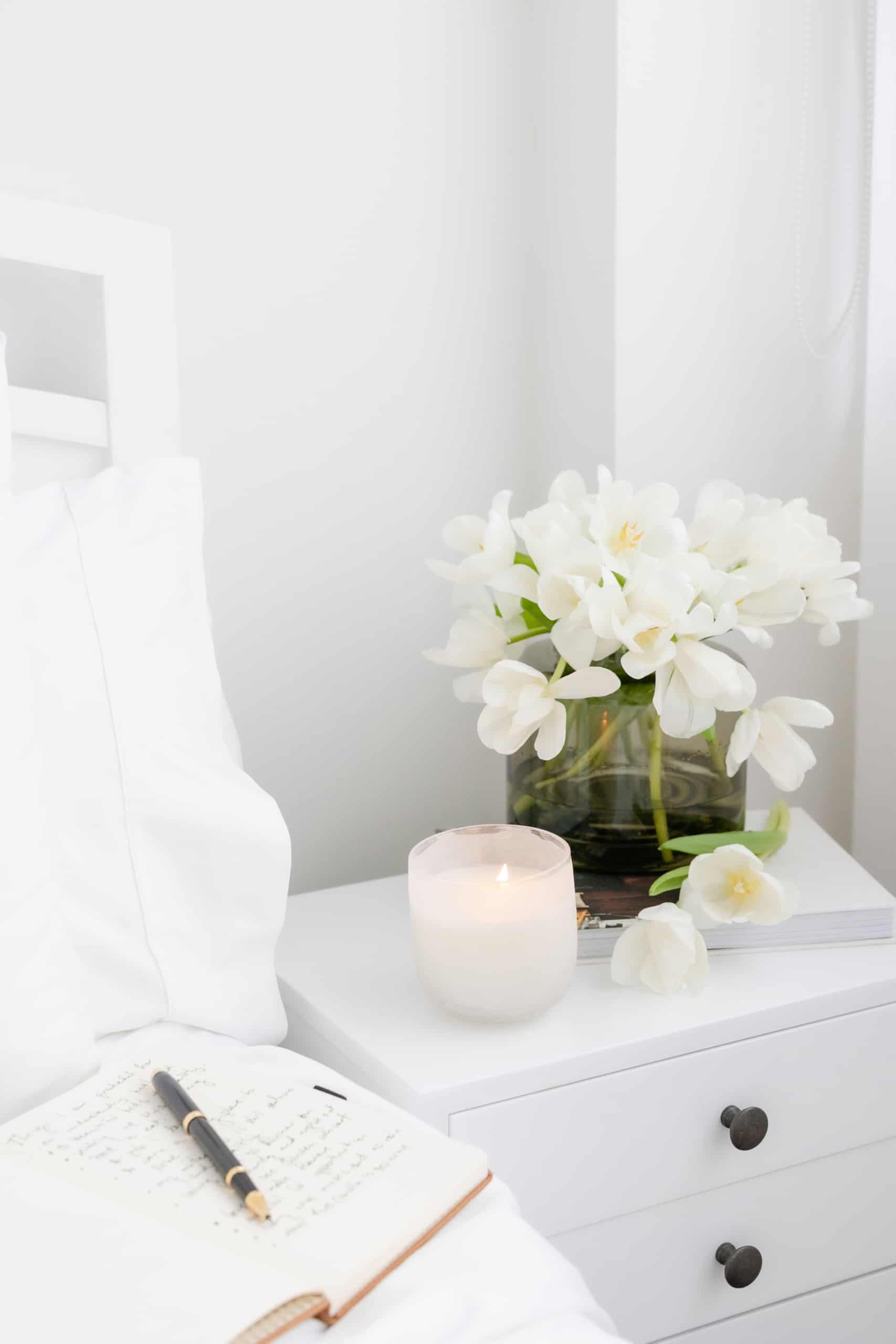 bedside table with flowers and candle next to bed with open journal and pen