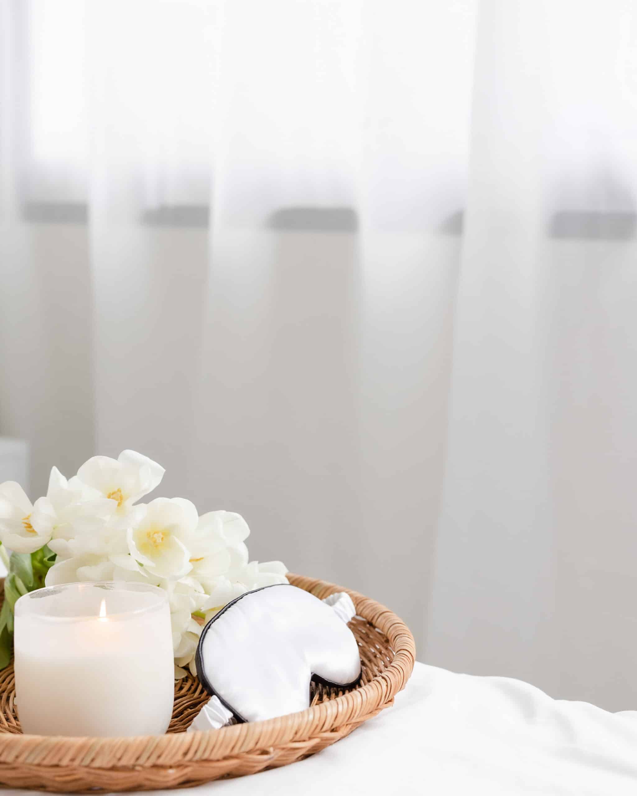 wicker tray on a bed that contains a burning candle, sleep mask, and fresh flowers. Light airy drapes are in the background