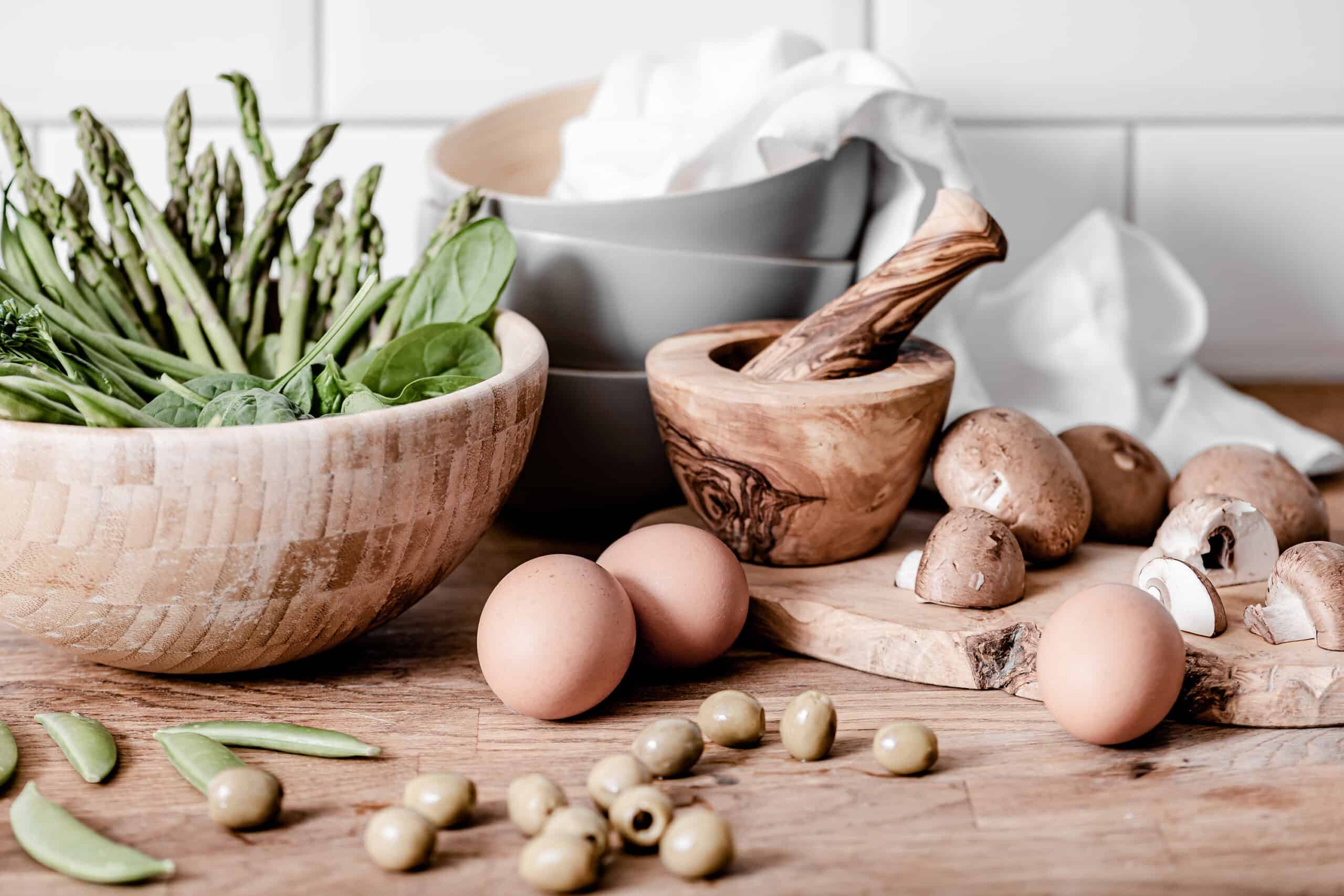 kitchen counter with a bowl filled with spinach & asparagus, eggs, olives, mushrooms, mortal and pestal, bowls, and cloth napkins - Copy