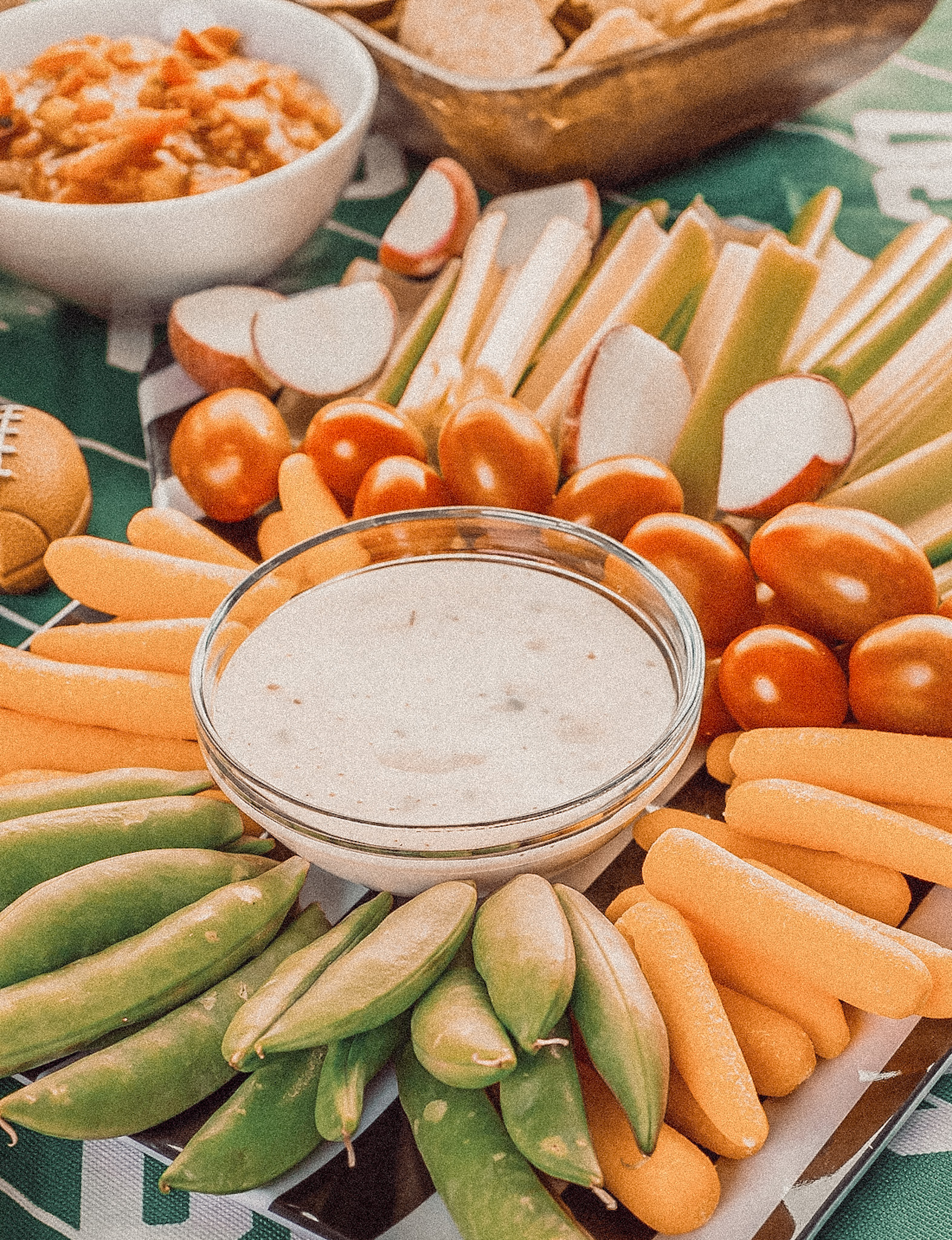 Overhead view of a veggie tray with dip, chips, and another dip on a football themed table.