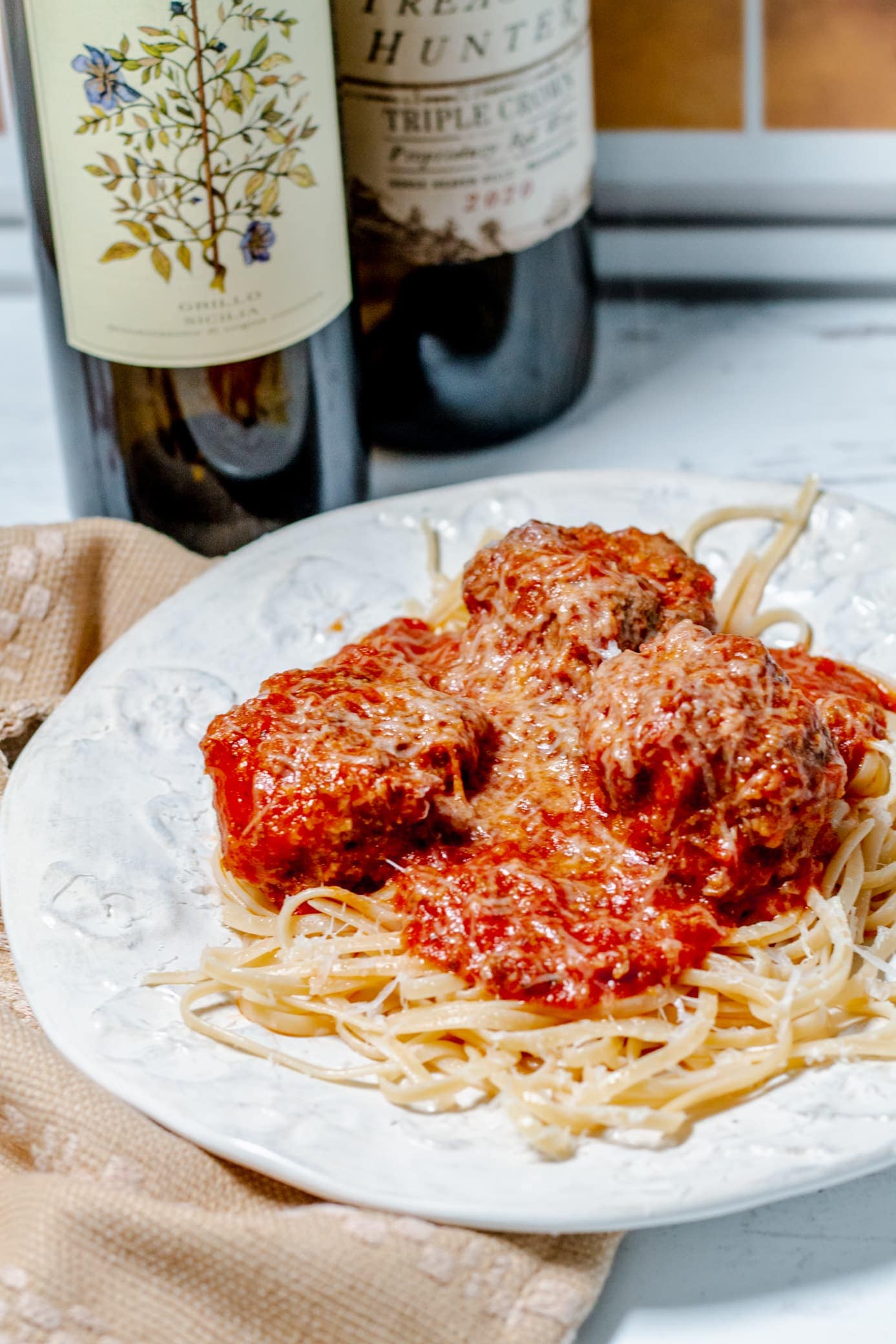 Classic spaghetti and meatballs on a white plate, made with all-ground beef meatballs and simmered in your favorite tomato sauce. The plate sits on a hand towel and there are 2 bottles of wine in the background.