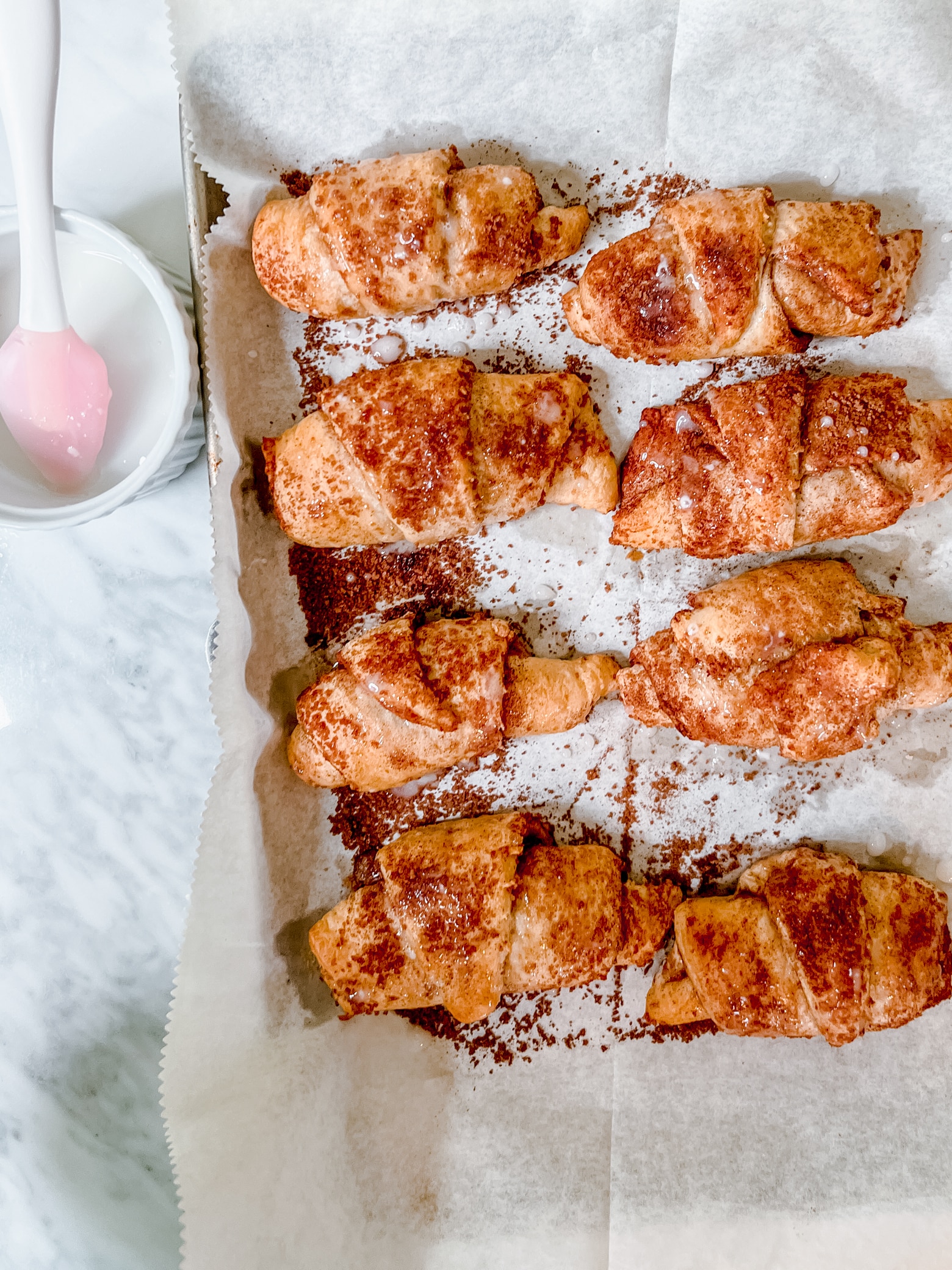 Yummy crescent cinnamon rolls on a baking sheet lined with parchment paper next to a baking spoon in a bowl.