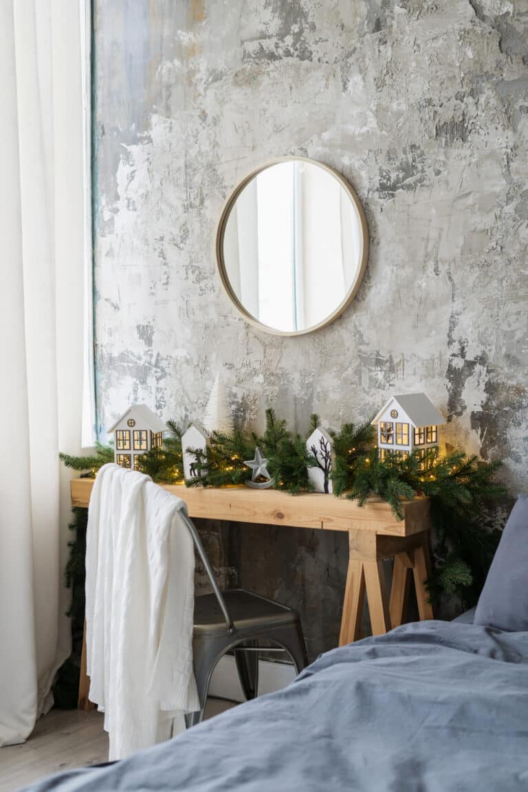 Vertical photo of cozy bedroom with loft style interior design, a circular mirror on the wall, wooden desk with green, winter decor on fir garland, chair with a soft white throw blanket sitting near a comfy looking bed.