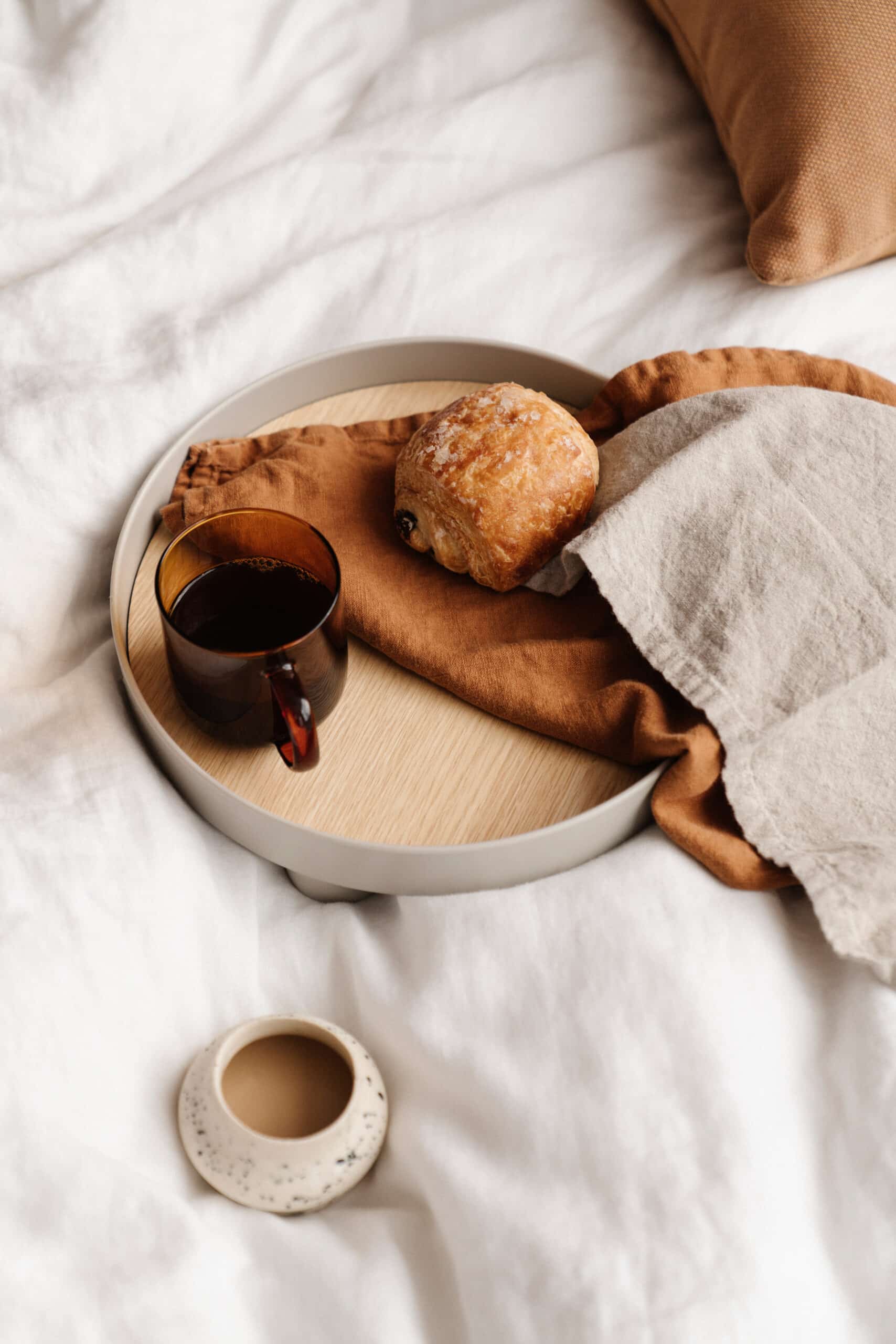 A tray is sitting on a comfy bed. The tray holds a cup of coffee, napkin, and pain au chocolate. There is an additional small cup next to the tray.