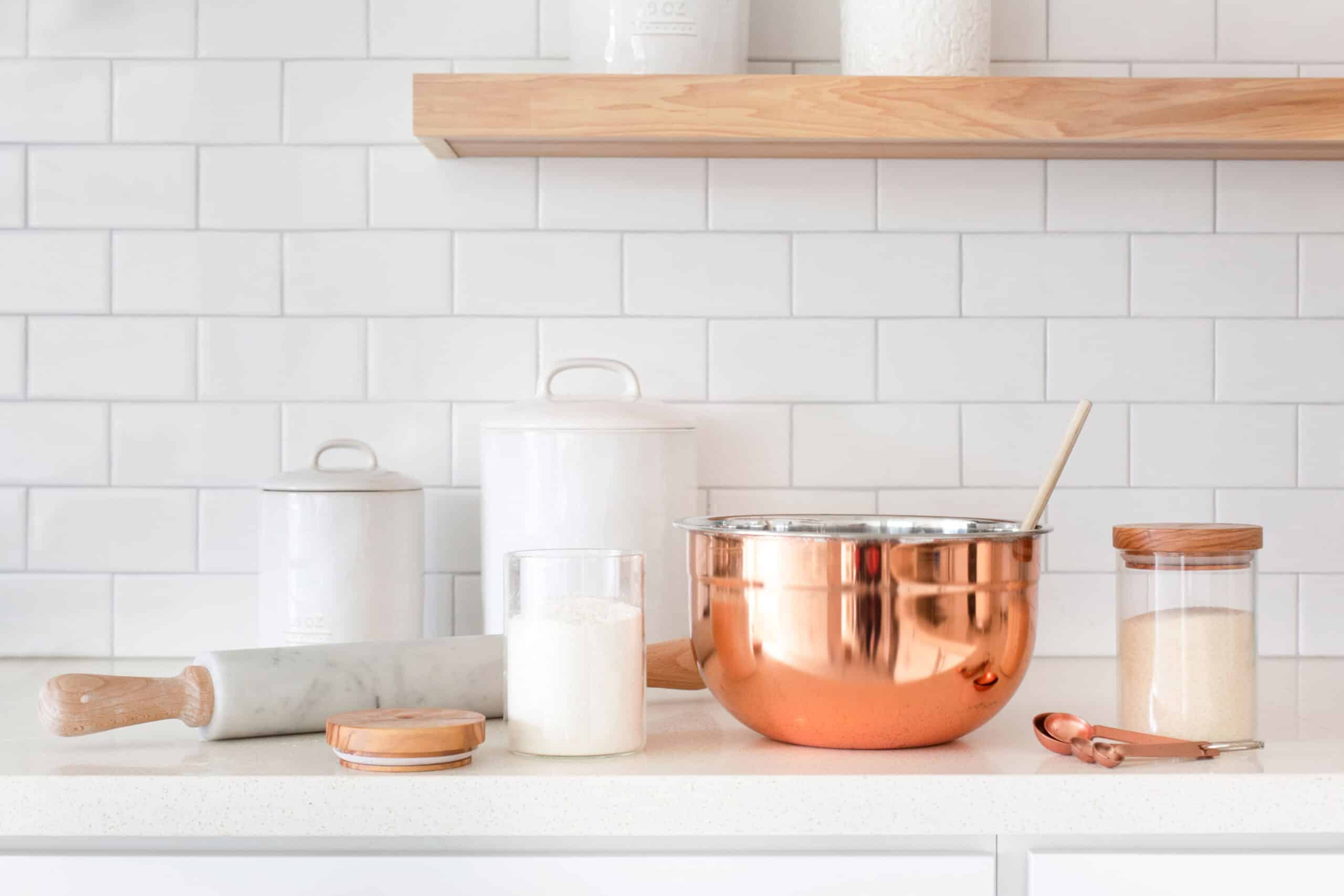 A kitchen counter laden with cooking supplies such as cannister for flour and sugar, a rolling pin, a metal bowl and wooden spoon, and measuring spoons.