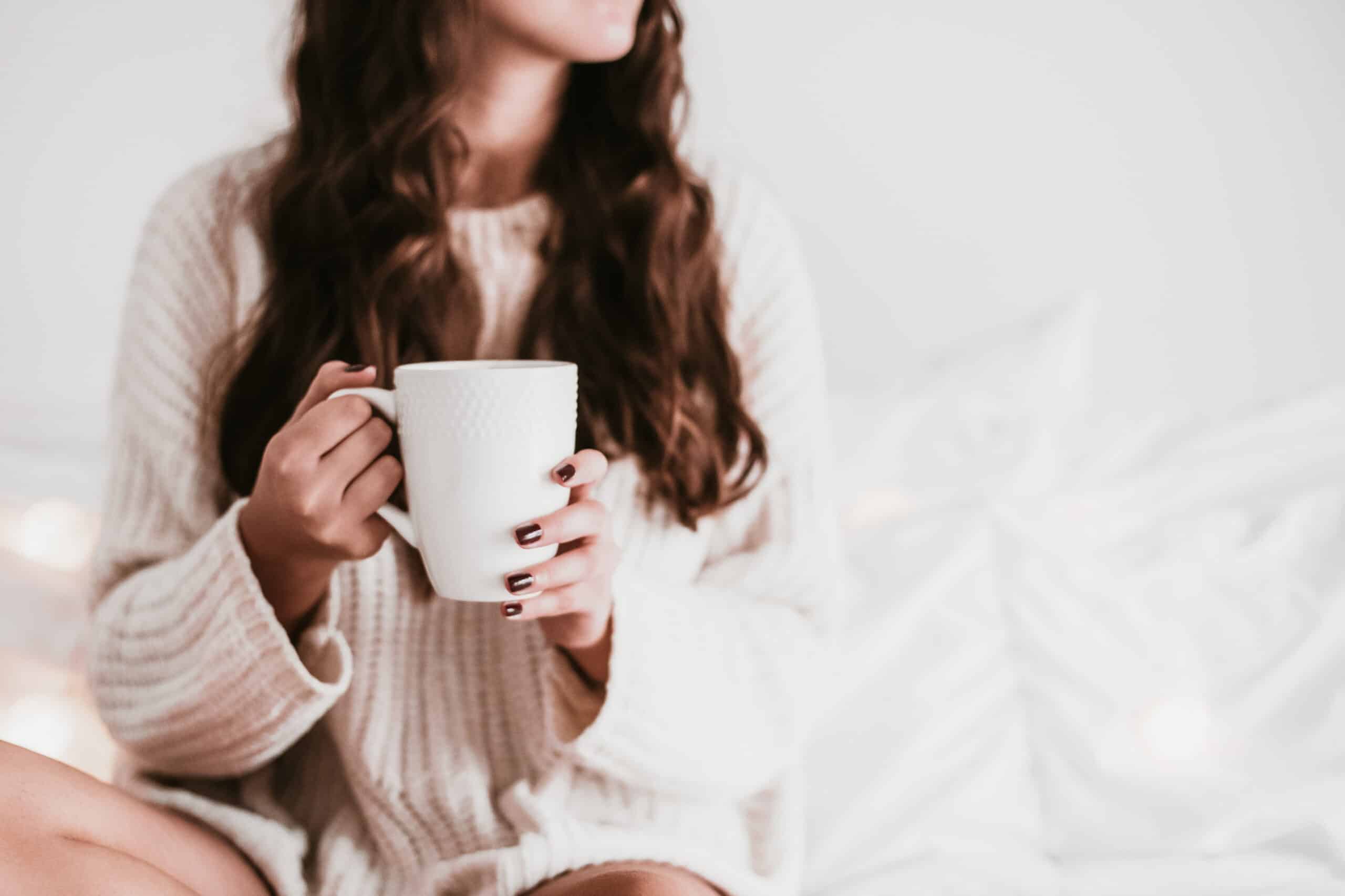 A woman sits on a comfy bed in a knit sweater holding a cup of tea.
