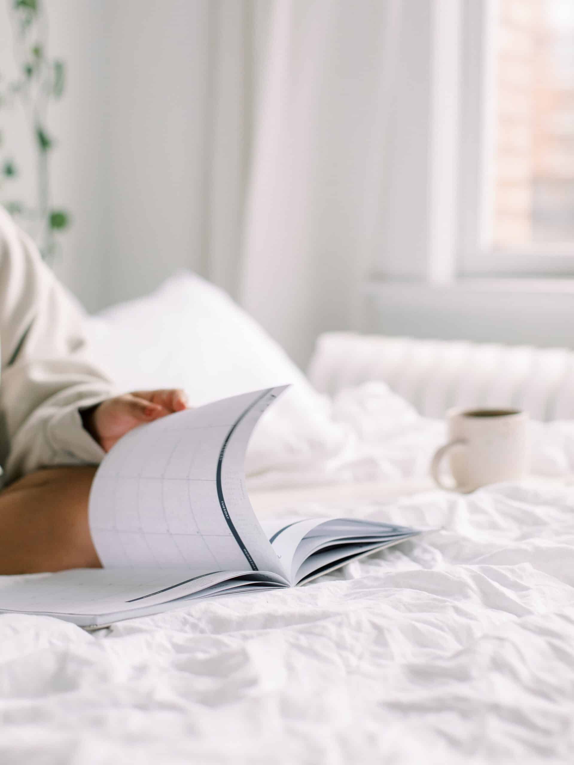 A person is flipping through the pages of a planner while they sit on a bed with a mug sitting next to them.