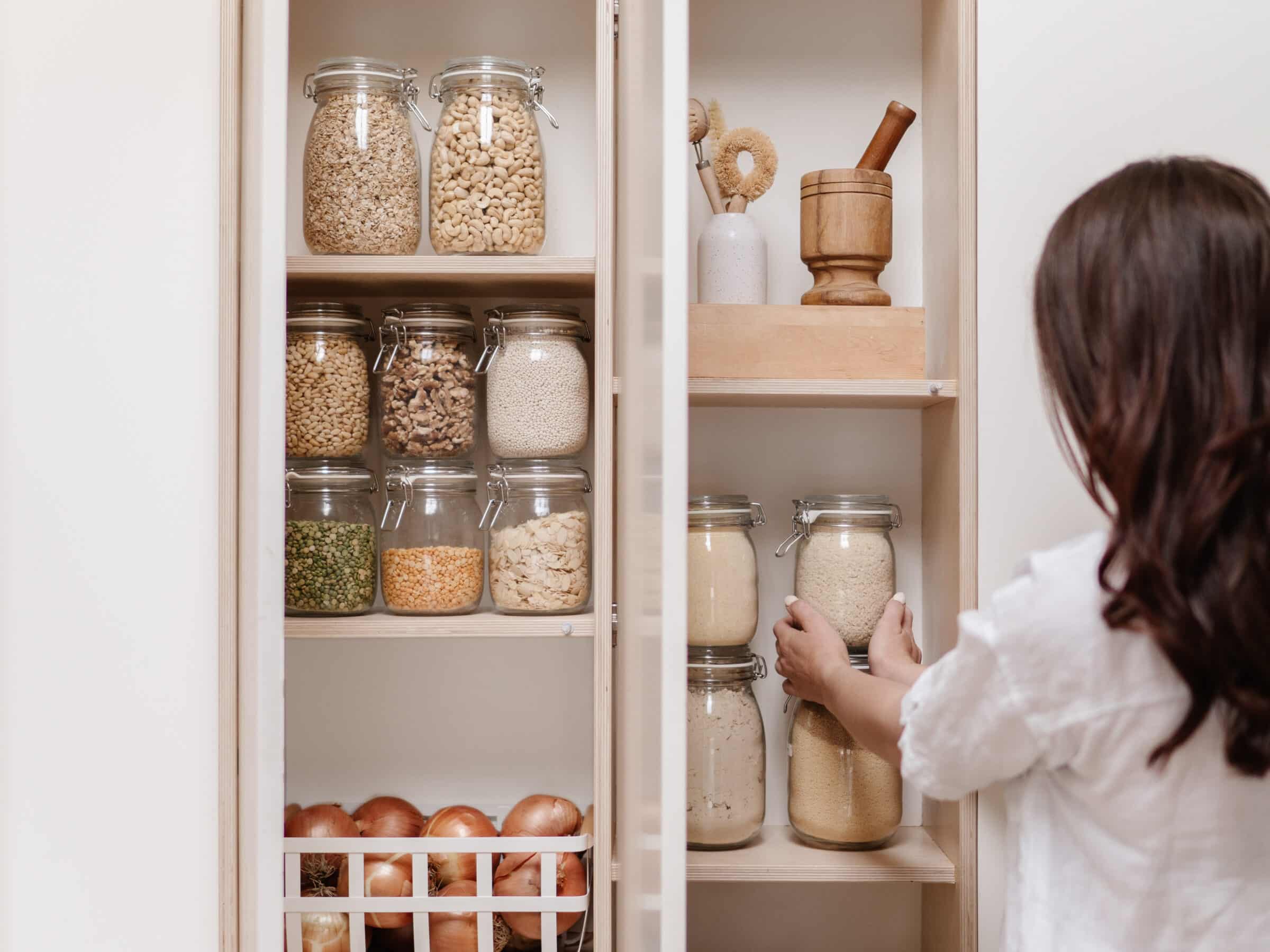 A woman places some glass jars filled with grains in a pantry. The rest of the pantry is filled with glass jars with other foods, a pestle and mortar, and some onions in a basket.