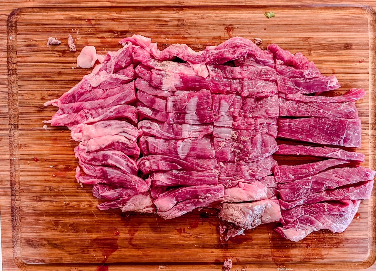 Steak cut into thin strips sits atop a wooden cutting board.