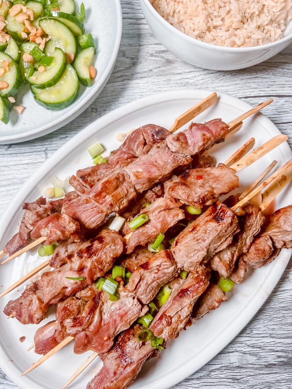 A table top holds plates and bowls full of cucumber salad, rice, and beef skewers seasoned with green onions.