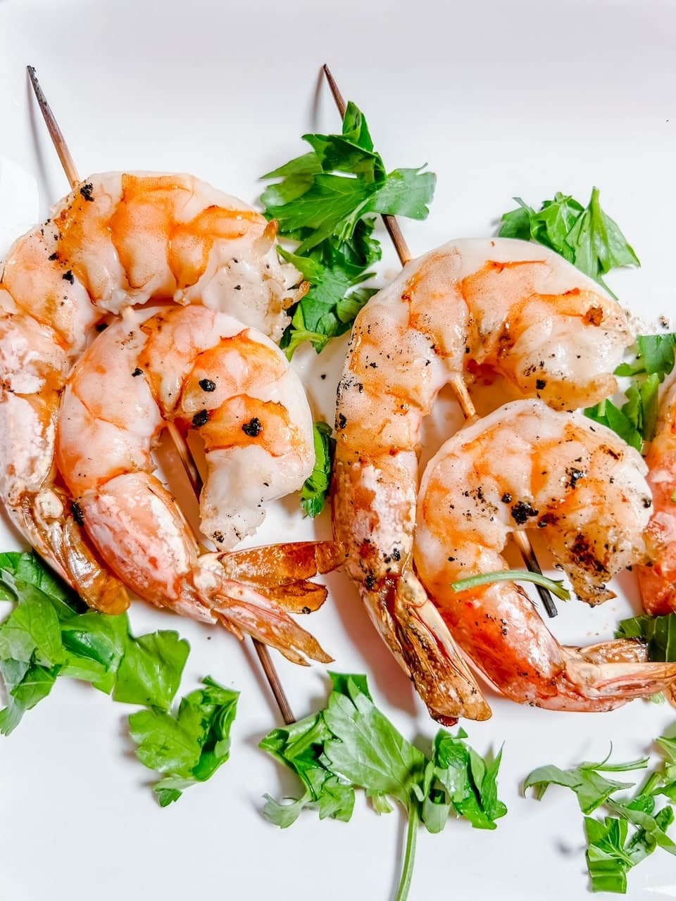 2 grilled shrimp skewers surrounded by a sprinkling of parsley lie on a white backdrop.