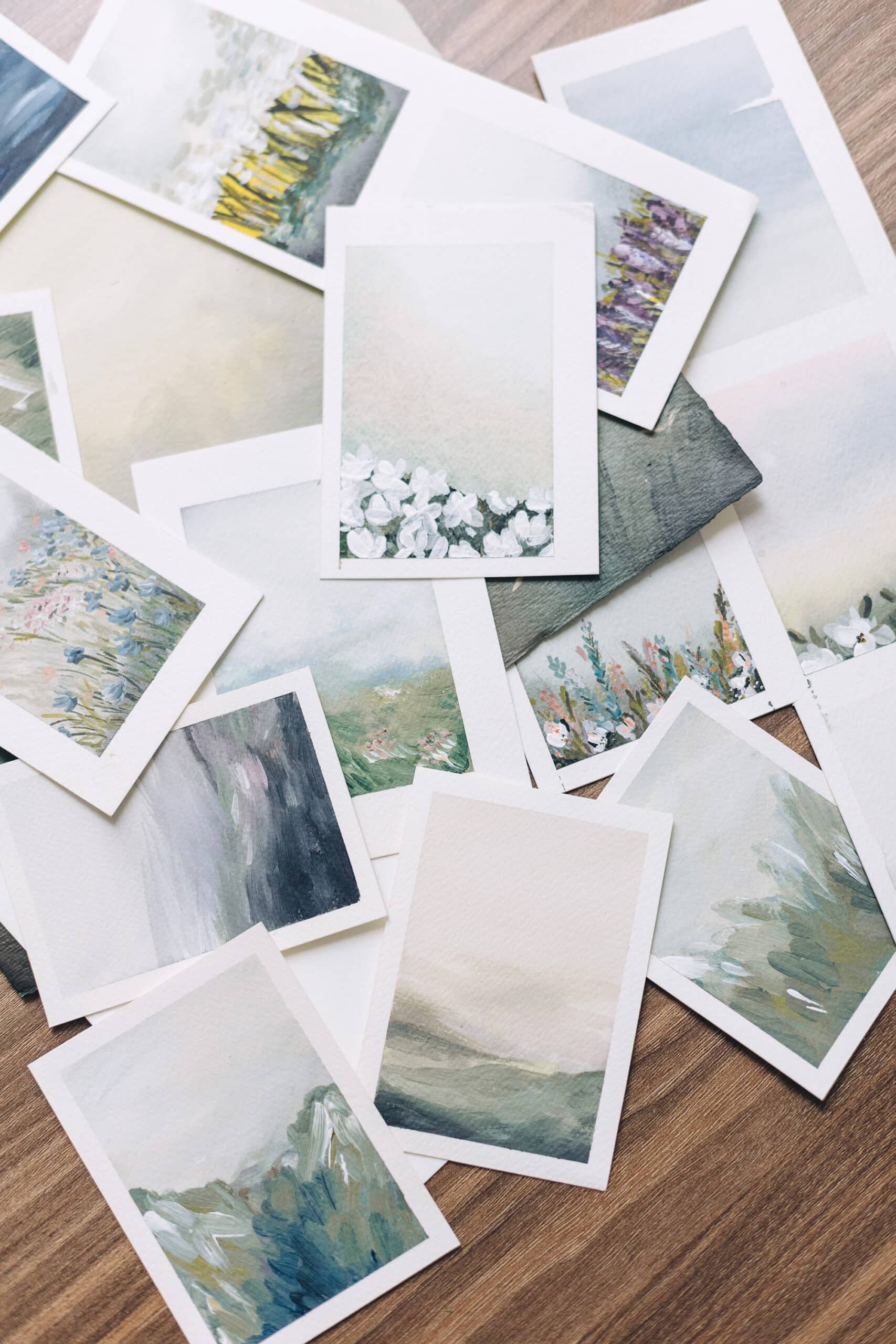 A pile of watercolor photos lies atop a wooden table. The images depict springtime foggy scenes.