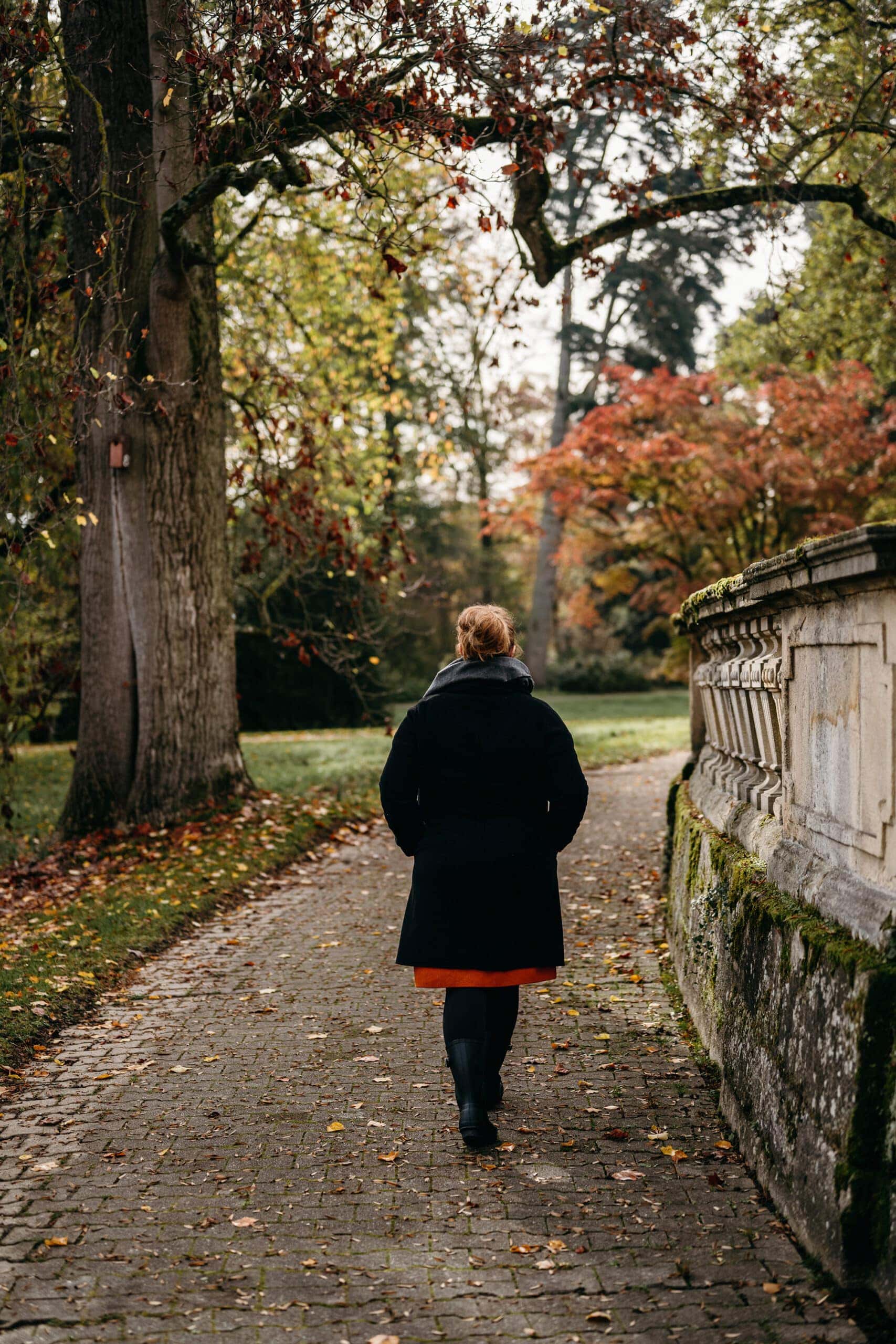 A woman walks away from view on a brick walkway with a stone wall to her right and a park with trees to her left.