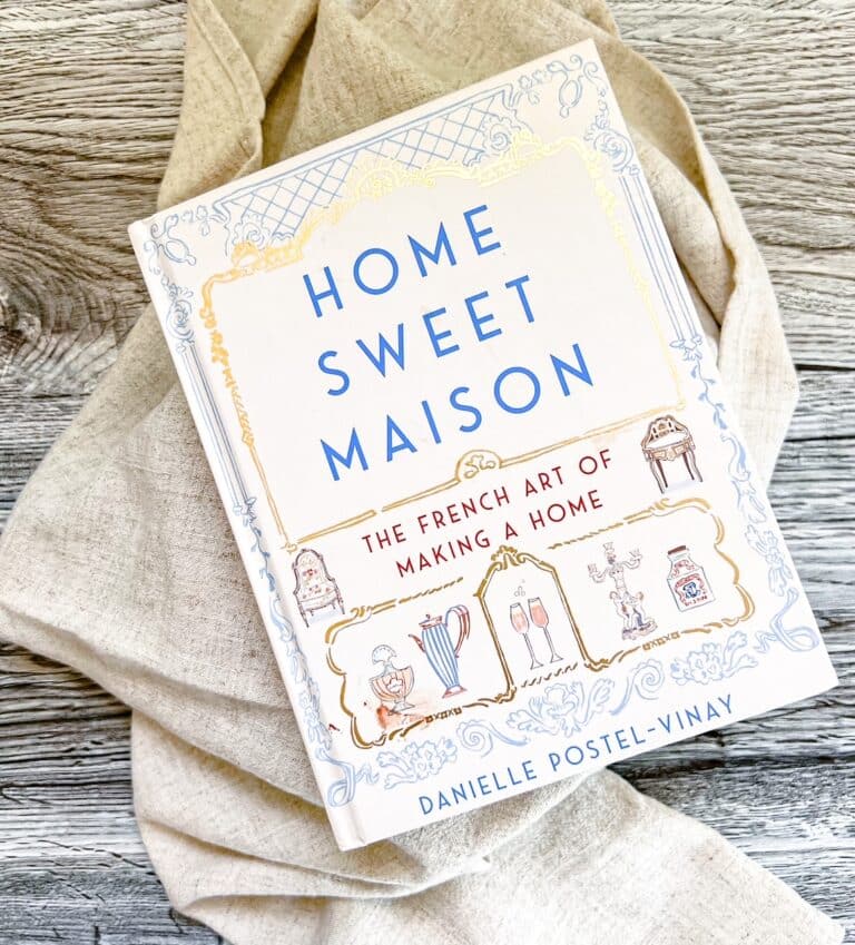 The book "Home Sweet Maison: The French Art of Making a Home" by Danielle Postel-Vinay sits on a linen cloth on a wooden tabletop.