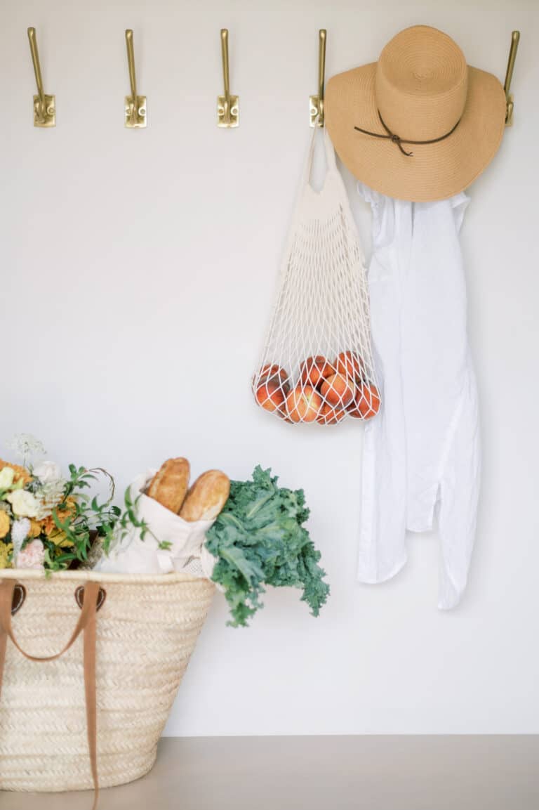 A shot of an entry way wall with hooks and a bench. On the left-hand side of the bench sits a tote bag filled with fresh flowers, bread, and vegetables. On the hooks on the right-hand side hang a bag of fruit, sunhat, and white button down shirt.