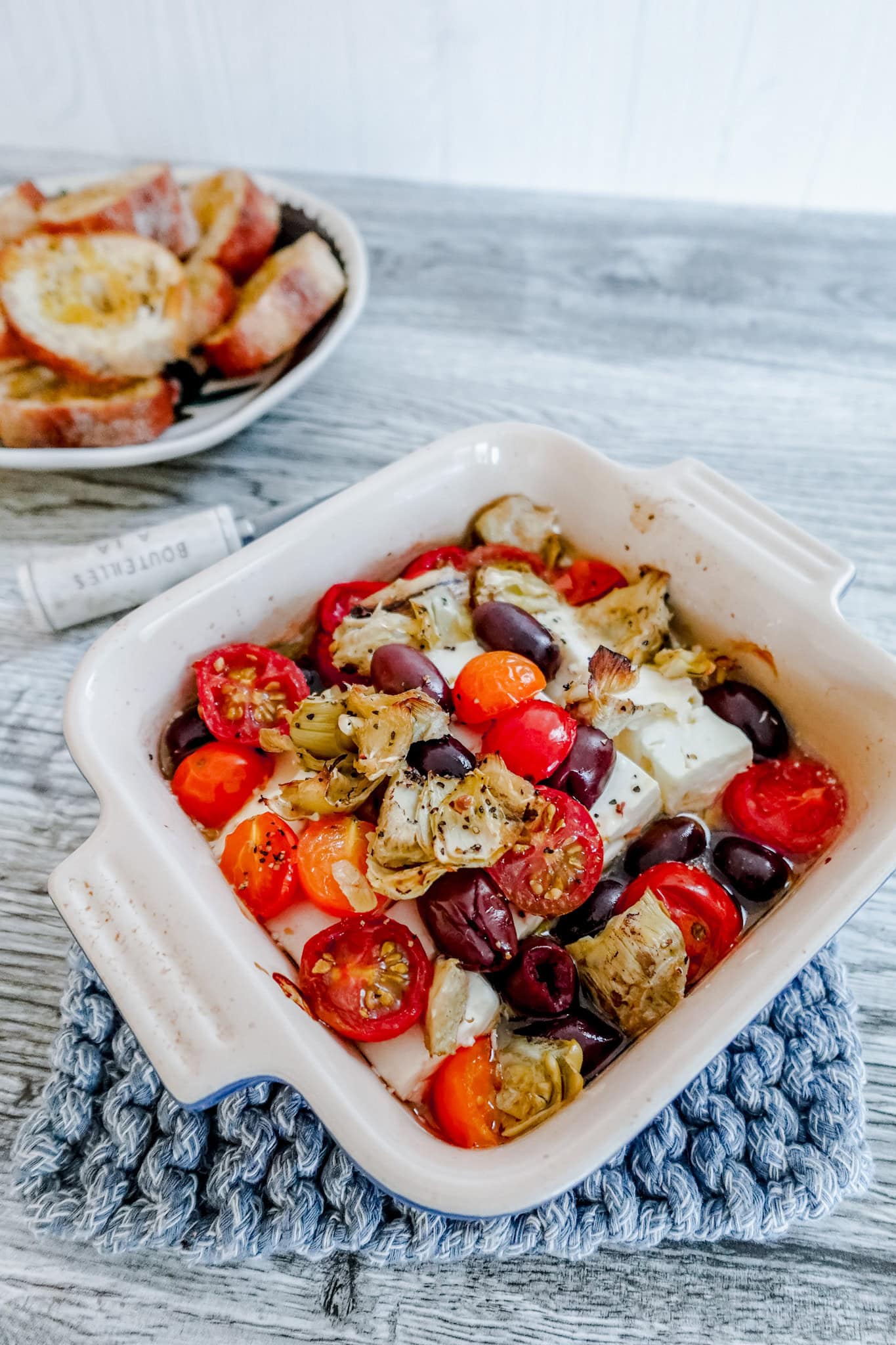 A white baking dish sits on a blue woven hot pad. In the dish is a baked feta cheese block, tomatoes, olives, and artichokes. There is a cheese knife and plate of crostini in the background.