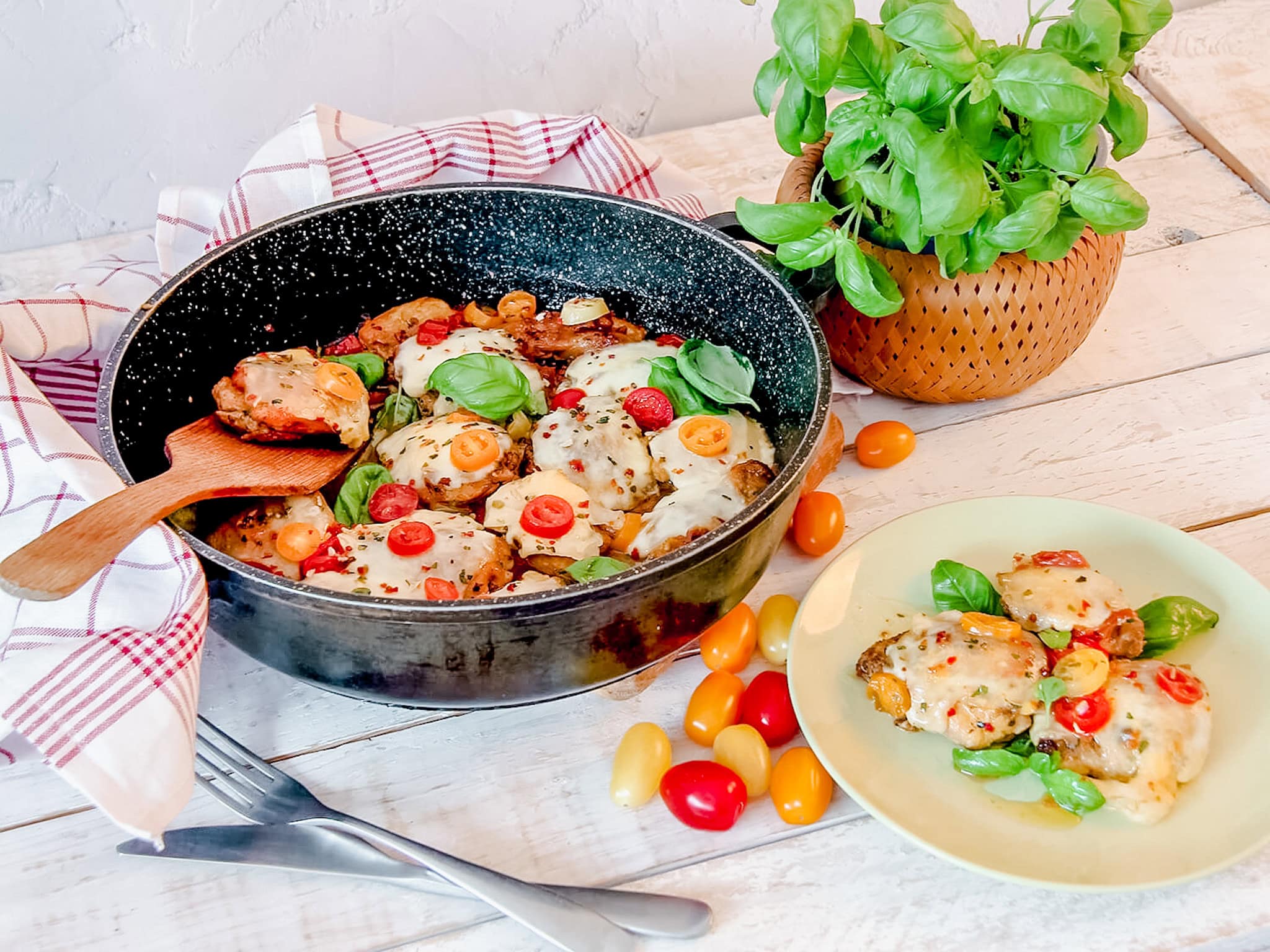 A full dinner spread featuring a skillet of Caprese Chicken Thighs alongside a plate with a serving of the dish. Fresh cherry tomatoes and a basil plant accent the rustic, cozy kitchen setting.
