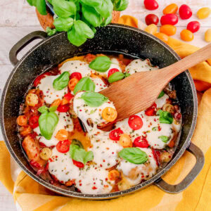 A skillet filled with Caprese Chicken Thighs, topped with melted mozzarella cheese, vibrant cherry tomatoes, and fresh basil leaves. A wooden spatula lifts one chicken thigh, highlighting its cheesy, juicy goodness. A yellow napkin and basil plant are visible in the background.