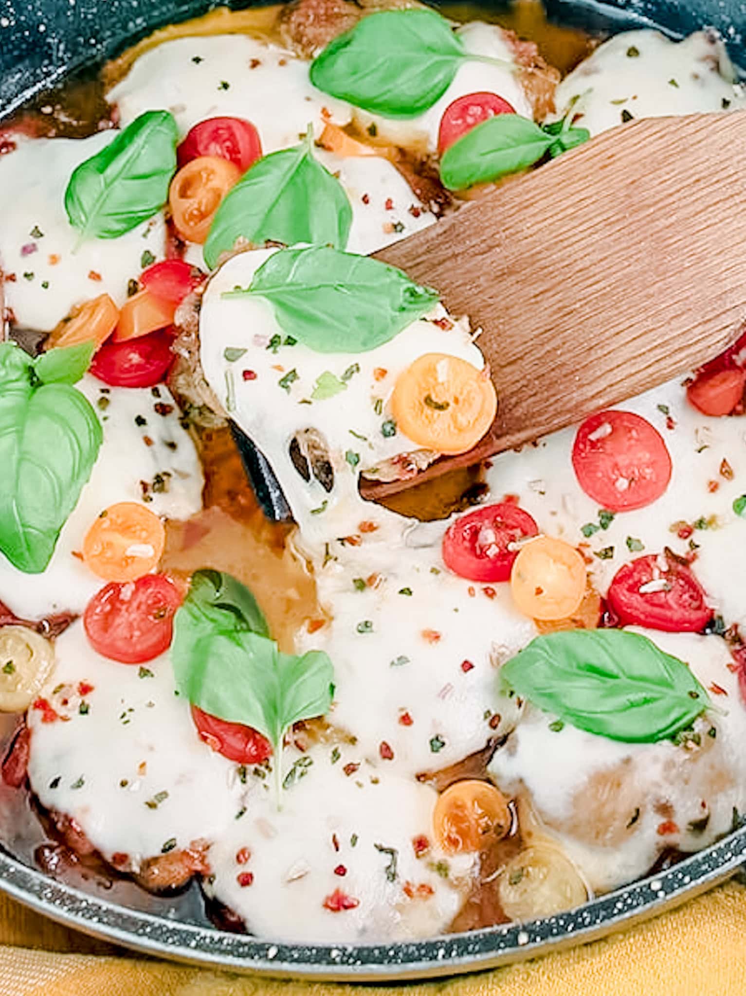 A close-up view of Caprese Chicken Thighs in a skillet, featuring golden-brown chicken thighs covered with melted cheese, sliced cherry tomatoes, and fresh basil leaves. A wooden spatula scoops up a piece, showcasing the delicious layers.