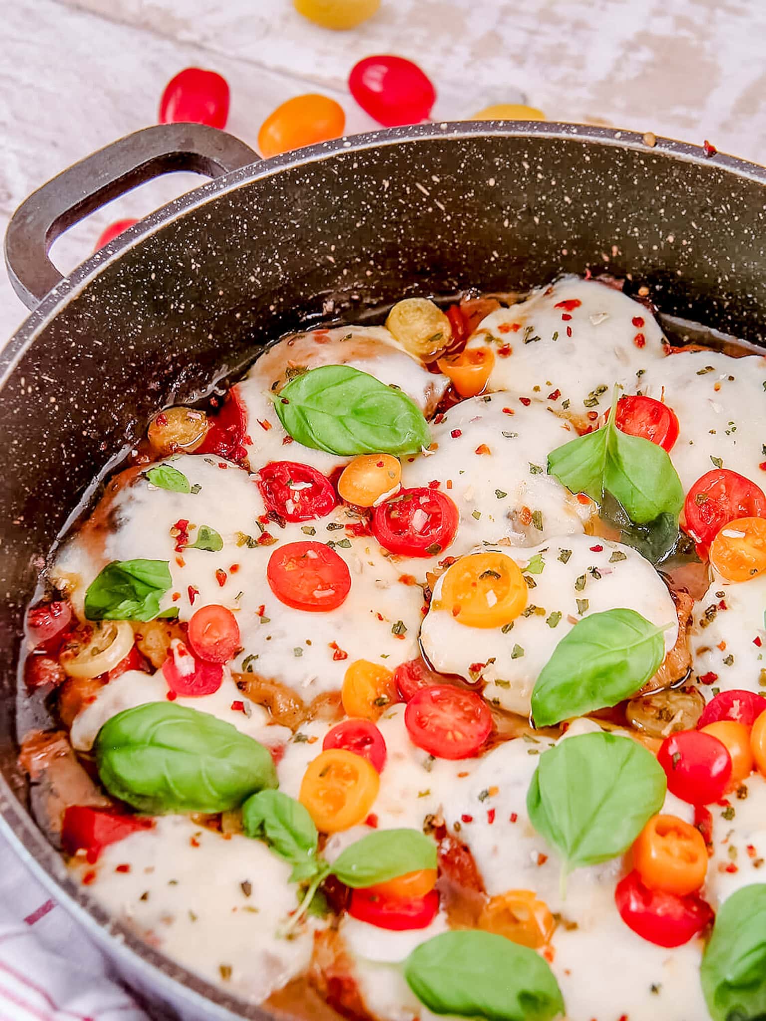A vibrant close-up of Caprese Chicken Thighs in a skillet, highlighting the melted mozzarella, colorful cherry tomato slices, and fresh basil leaves.
