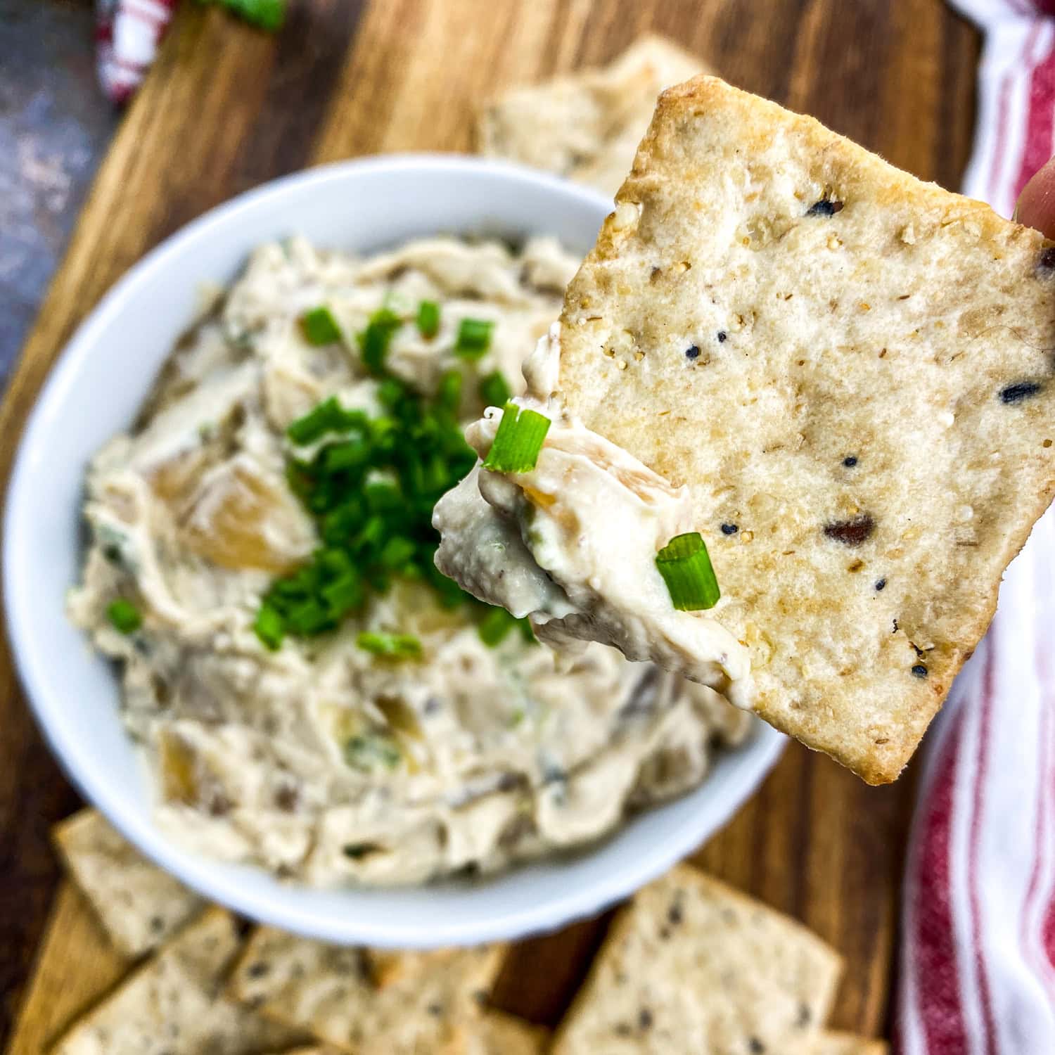A square photo of caramelized onion dip in a white bowl topped with chives, with a cracker dipped into the creamy dip, surrounded by crackers and fresh vegetables.