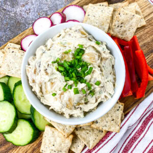A bowl of caramelized onion dip garnished with chives, set on a wooden serving board with crackers, sliced cucumbers, radishes, and red bell pepper arranged around it.