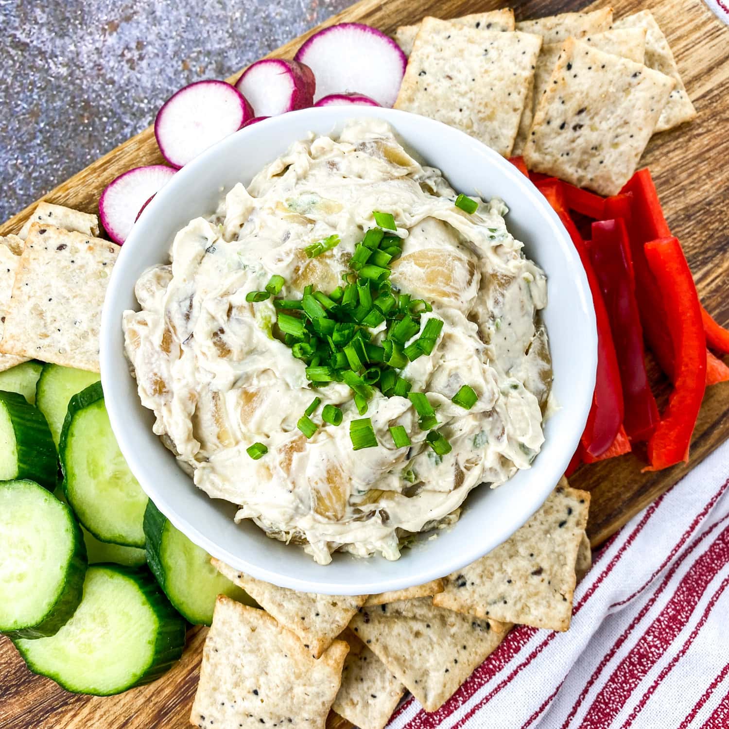 A bowl of caramelized onion dip garnished with chives, set on a wooden serving board with crackers, sliced cucumbers, radishes, and red bell pepper arranged around it.