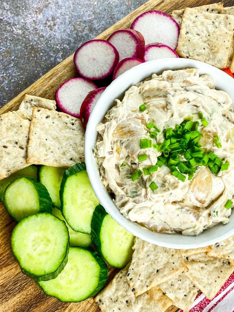 A close-up of caramelized onion dip in a white bowl, topped with fresh chopped chives, served with assorted crackers, sliced cucumbers, radishes, and red bell pepper on a wooden platter.