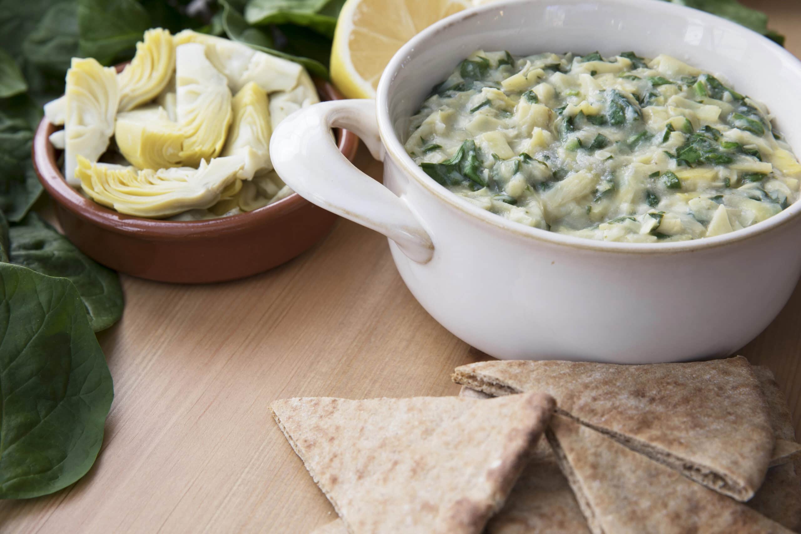 A creamy spinach and artichoke dip served in a white bowl, accompanied by a bowl of sliced artichokes and fresh spinach leaves, with pita bread wedges on the side.